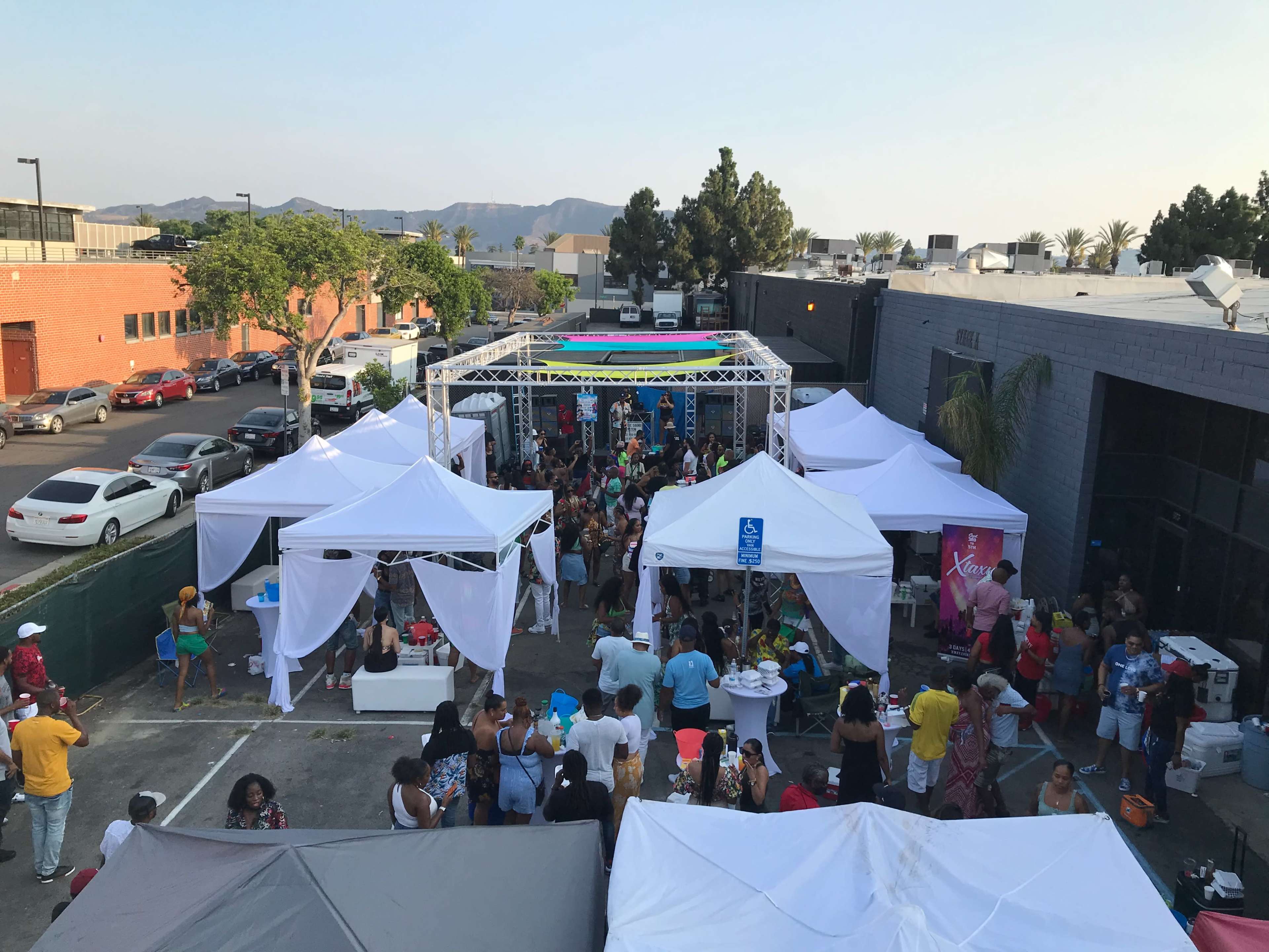 A large outdoor event with multiple white tents and a crowd of people gathered in a parking lot, surrounded by commercial buildings and palm trees.