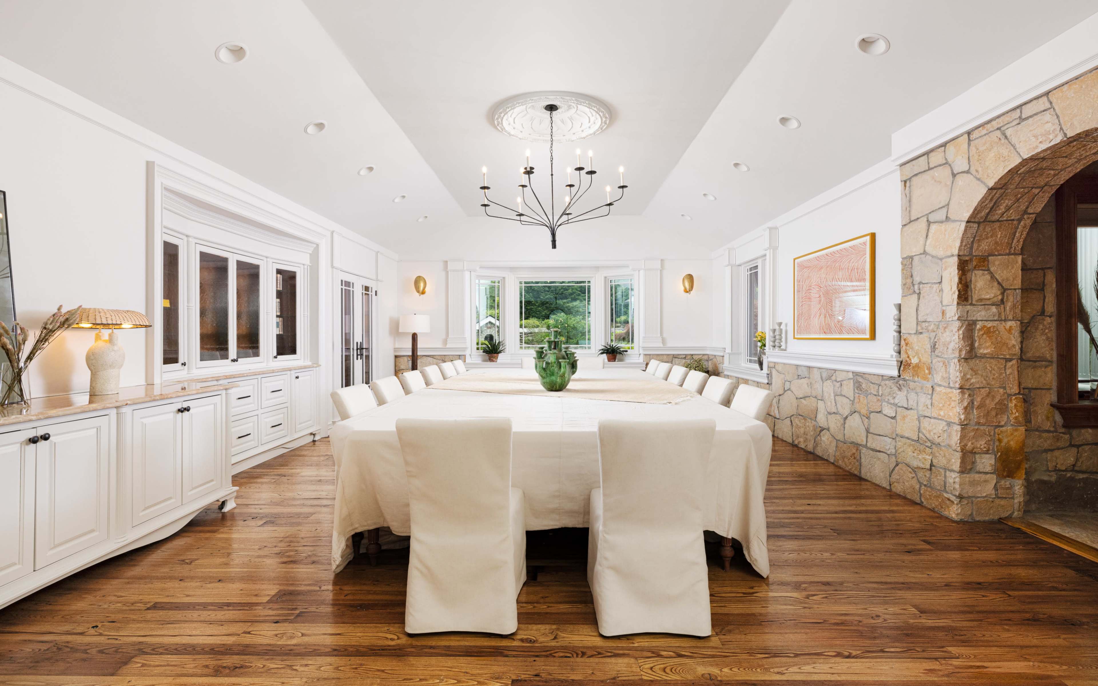 A spacious dining room features a long table surrounded by upholstered chairs, large windows, and a stone archway.