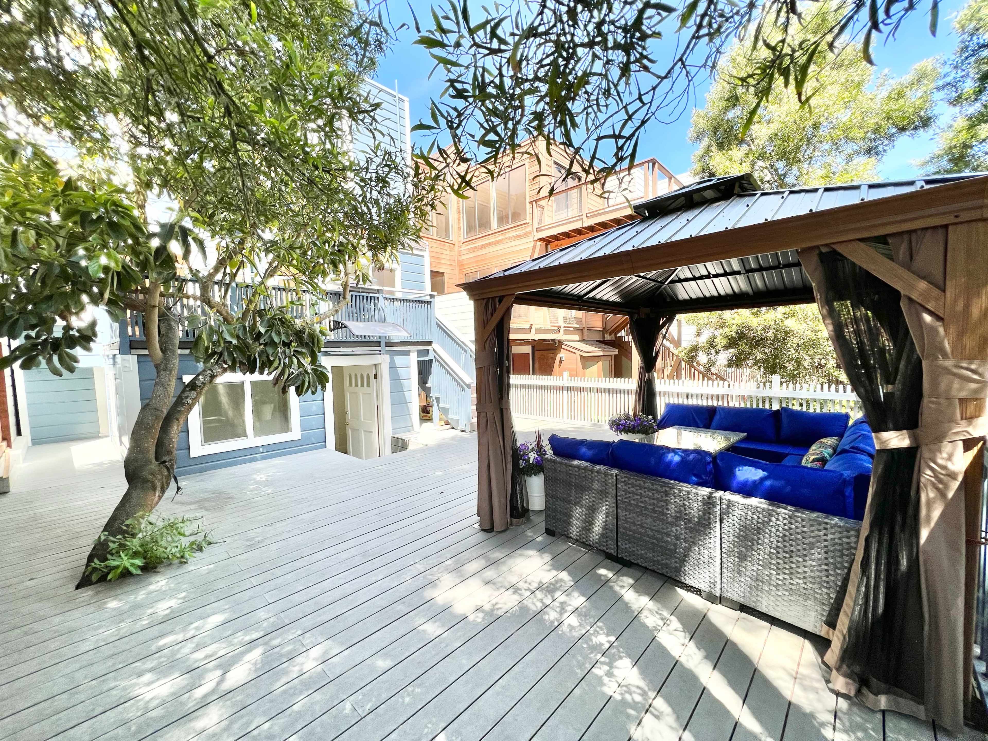A wooden deck features a gazebo with seating, surrounded by trees and adjacent to a multi-story building.