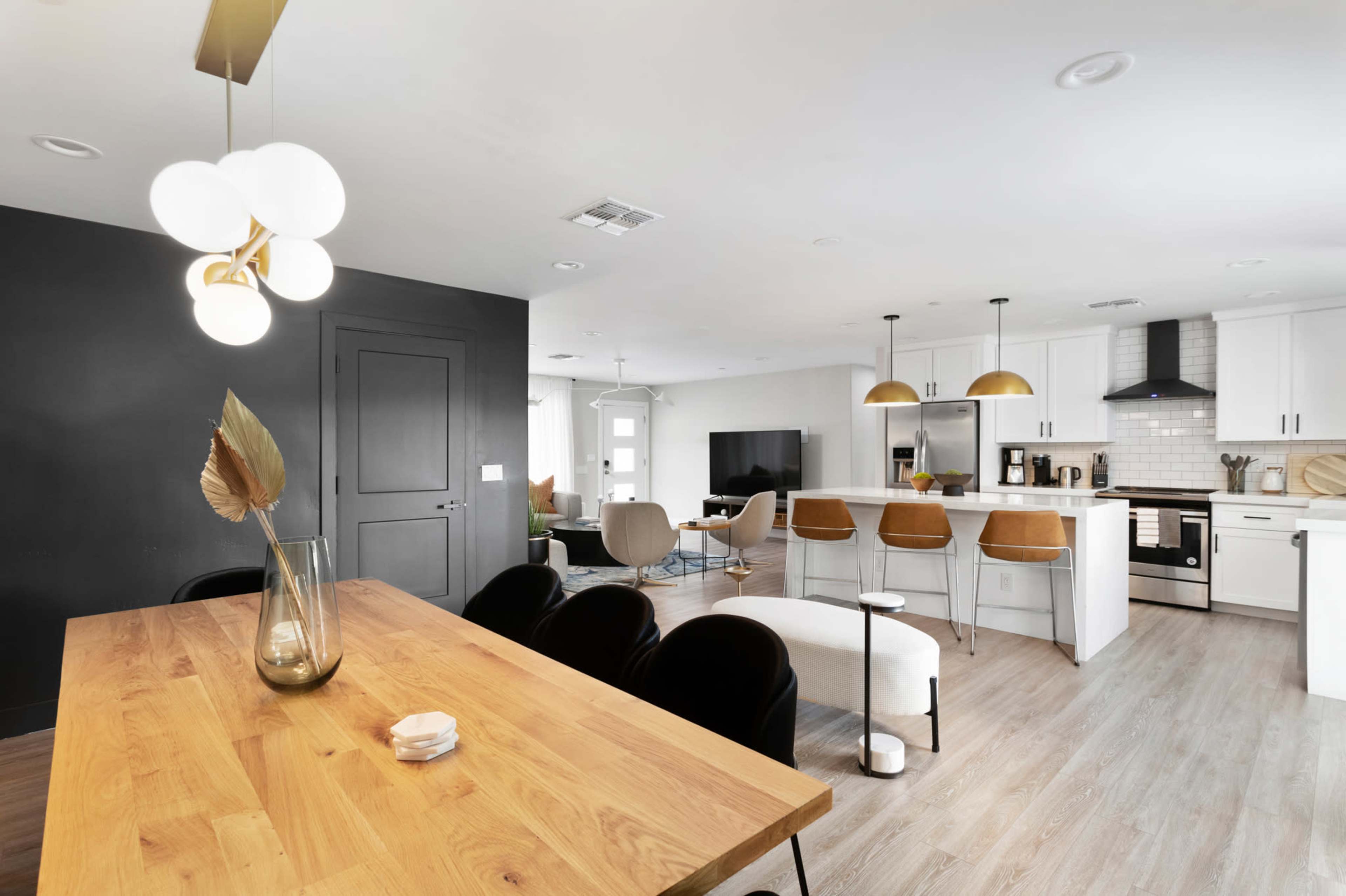 A modern open-concept living space featuring a dining area with a wooden table, a black accent wall, and a kitchen equipped with white cabinetry and gold pendant lights.