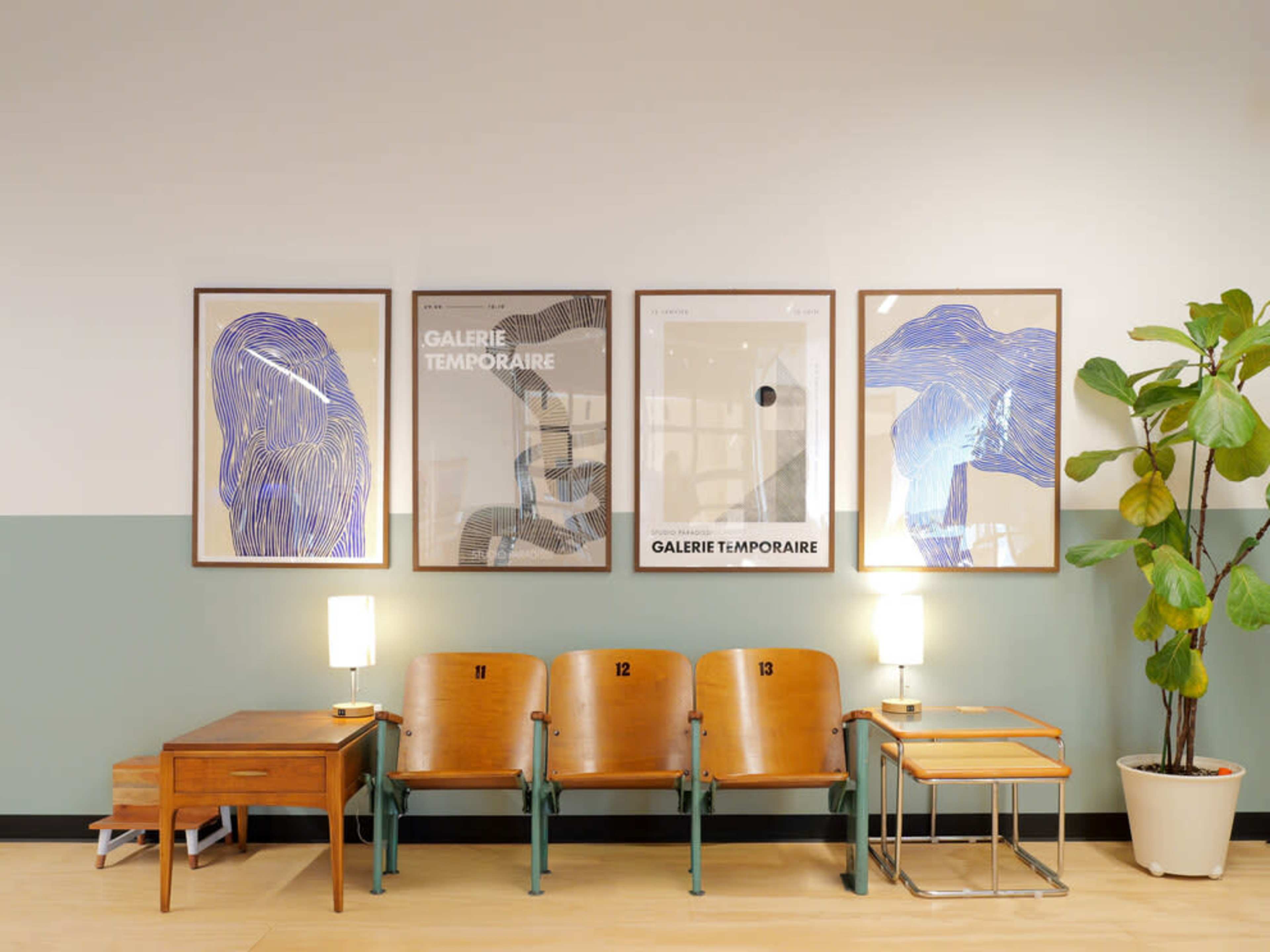 The image shows a waiting area featuring a row of wooden seating with three framed art pieces on the wall and a potted plant in the corner.