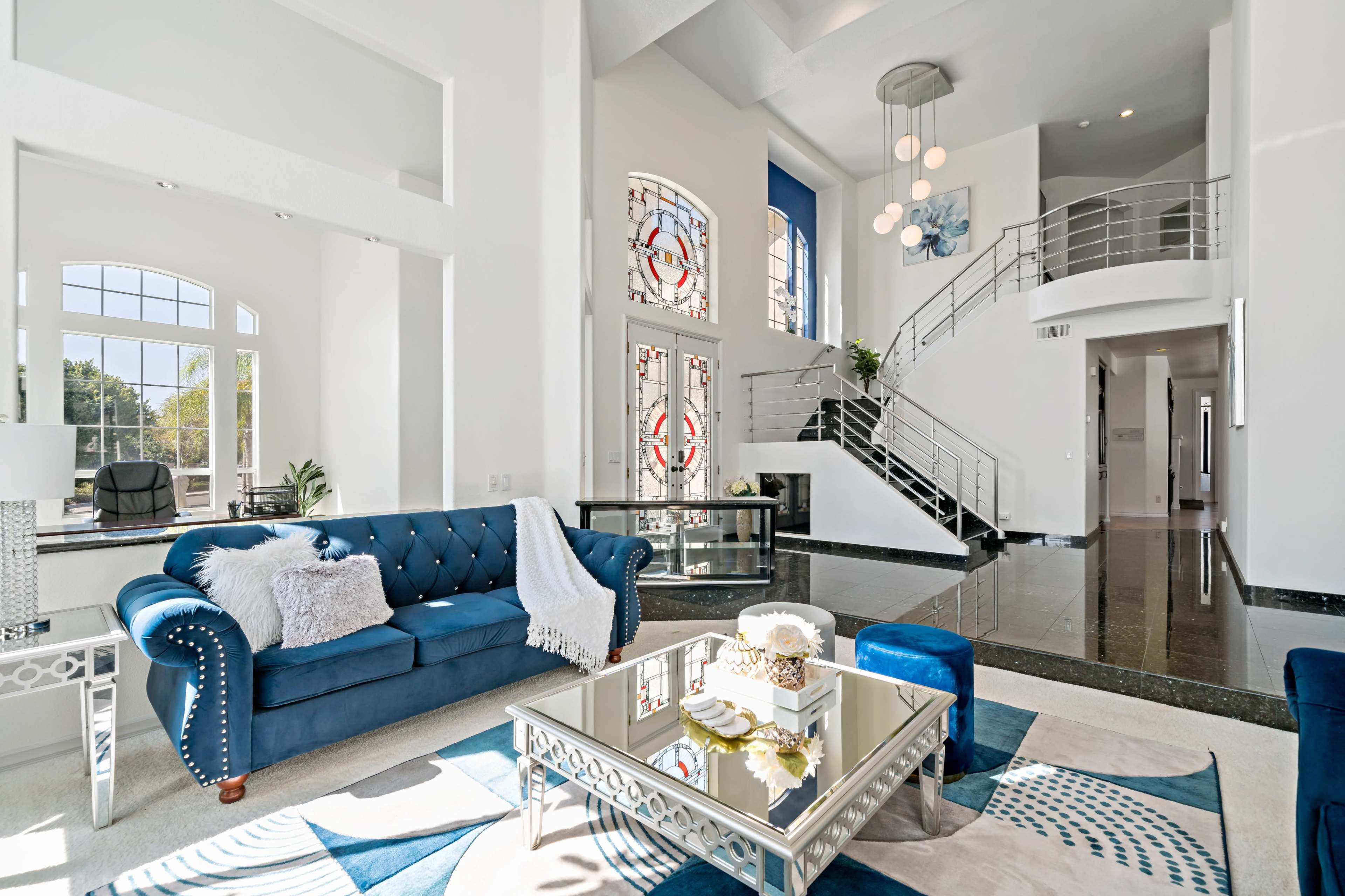A modern living room with a blue velvet sofa, a glass coffee table, and large windows that allow natural light to illuminate the space.