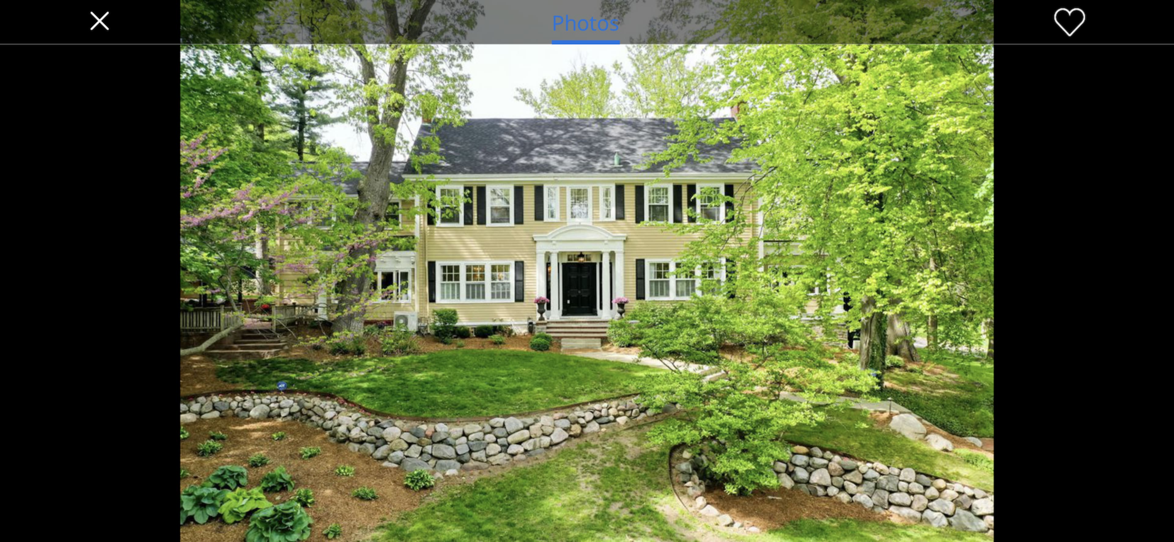 A two-story yellow house with a black roof, situated among lush green trees and a landscaped yard featuring a stone wall.