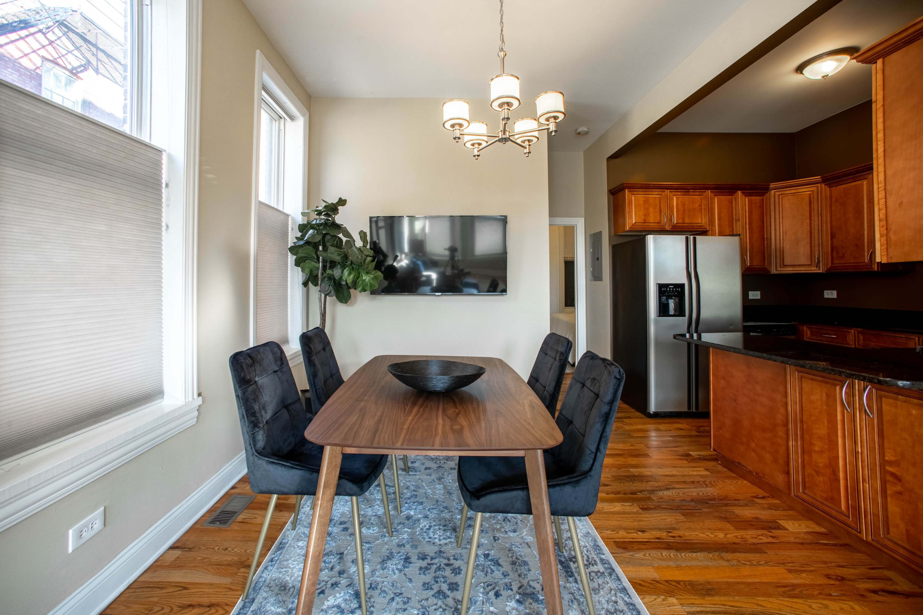 A dining area features a wooden table surrounded by four chairs, with a wall-mounted television and a plant nearby, illuminated by a chandelier above.