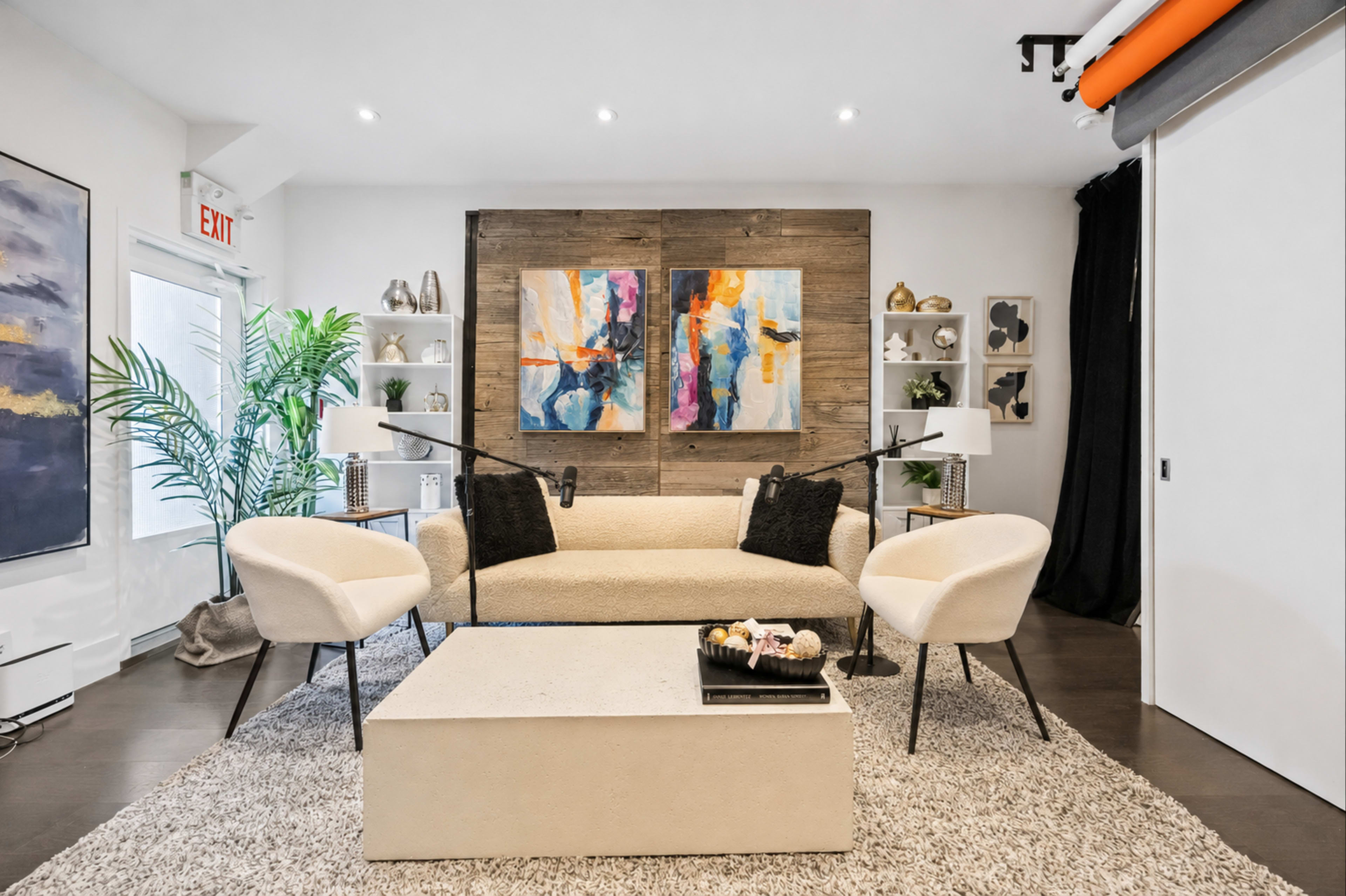 The image shows a modern living room featuring a cream-colored sofa, two light-colored chairs, a coffee table, and colorful abstract artwork on the wall, with potted plants and decorative shelves in the background.