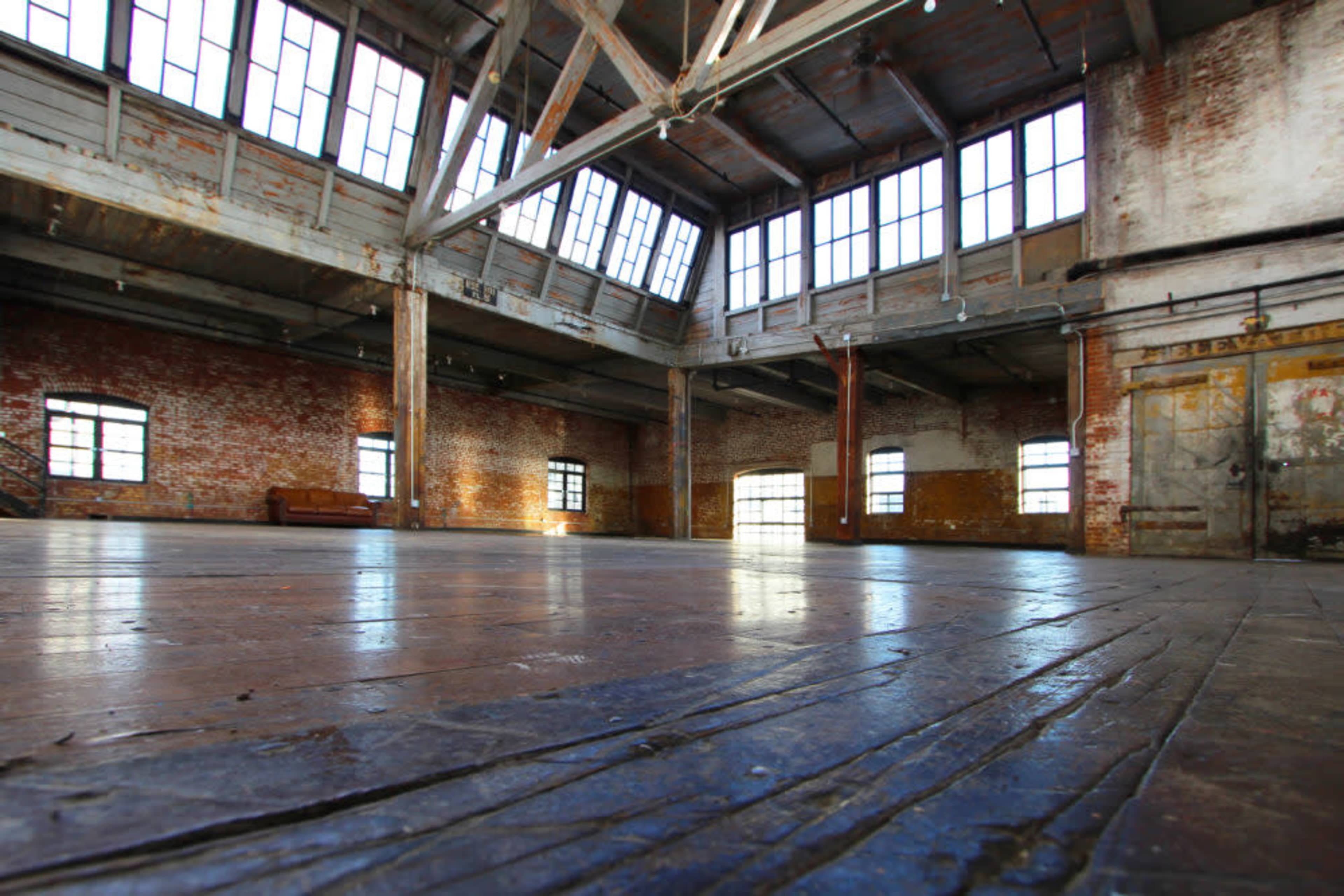 The image shows a spacious, industrial-style room with high ceilings, large windows, and exposed brick walls.
