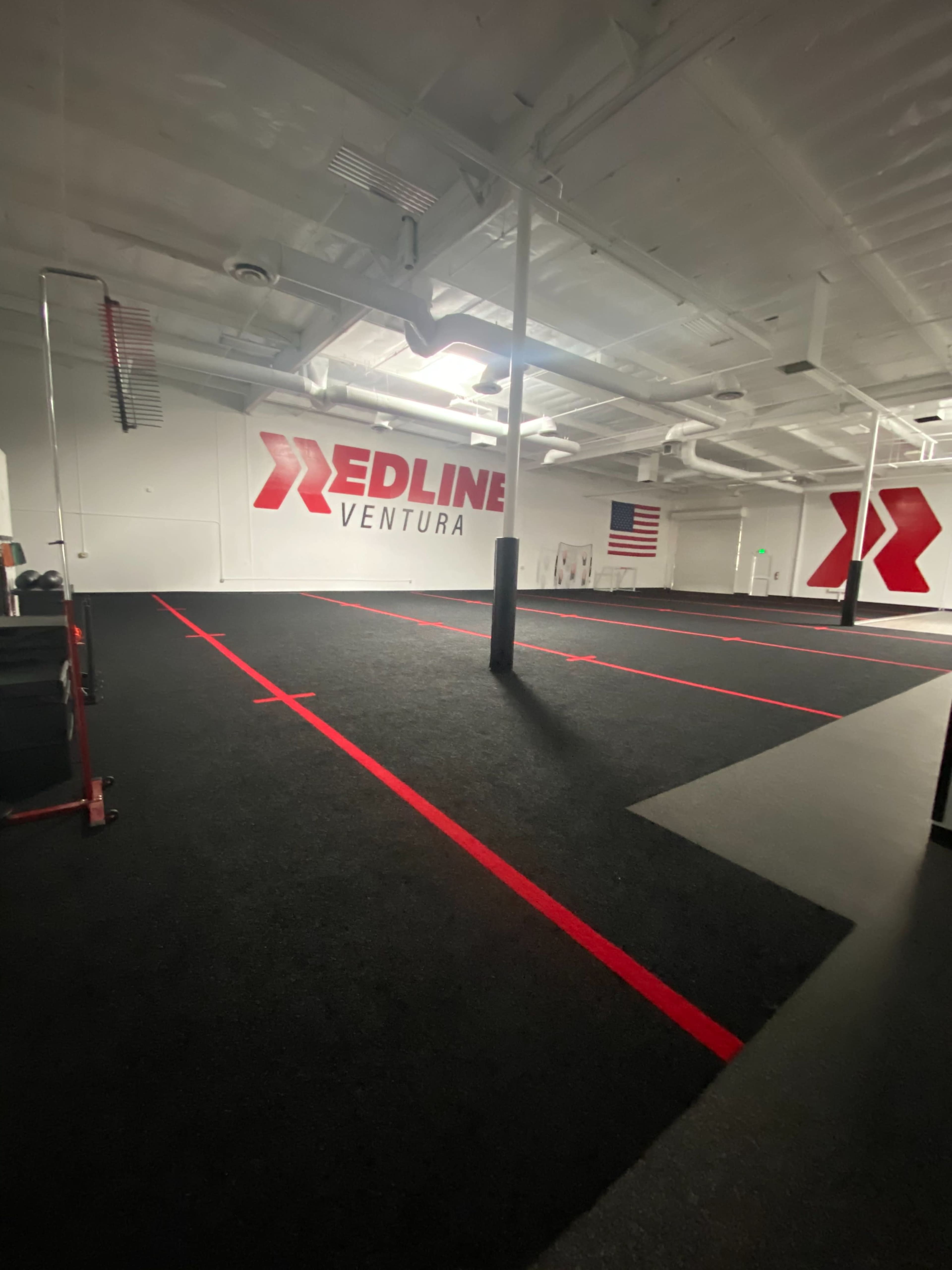 The image shows a spacious, equipped gym with black flooring, red lines marking workout areas, and a large wall displaying the "Redline Ventura" logo and an American flag.