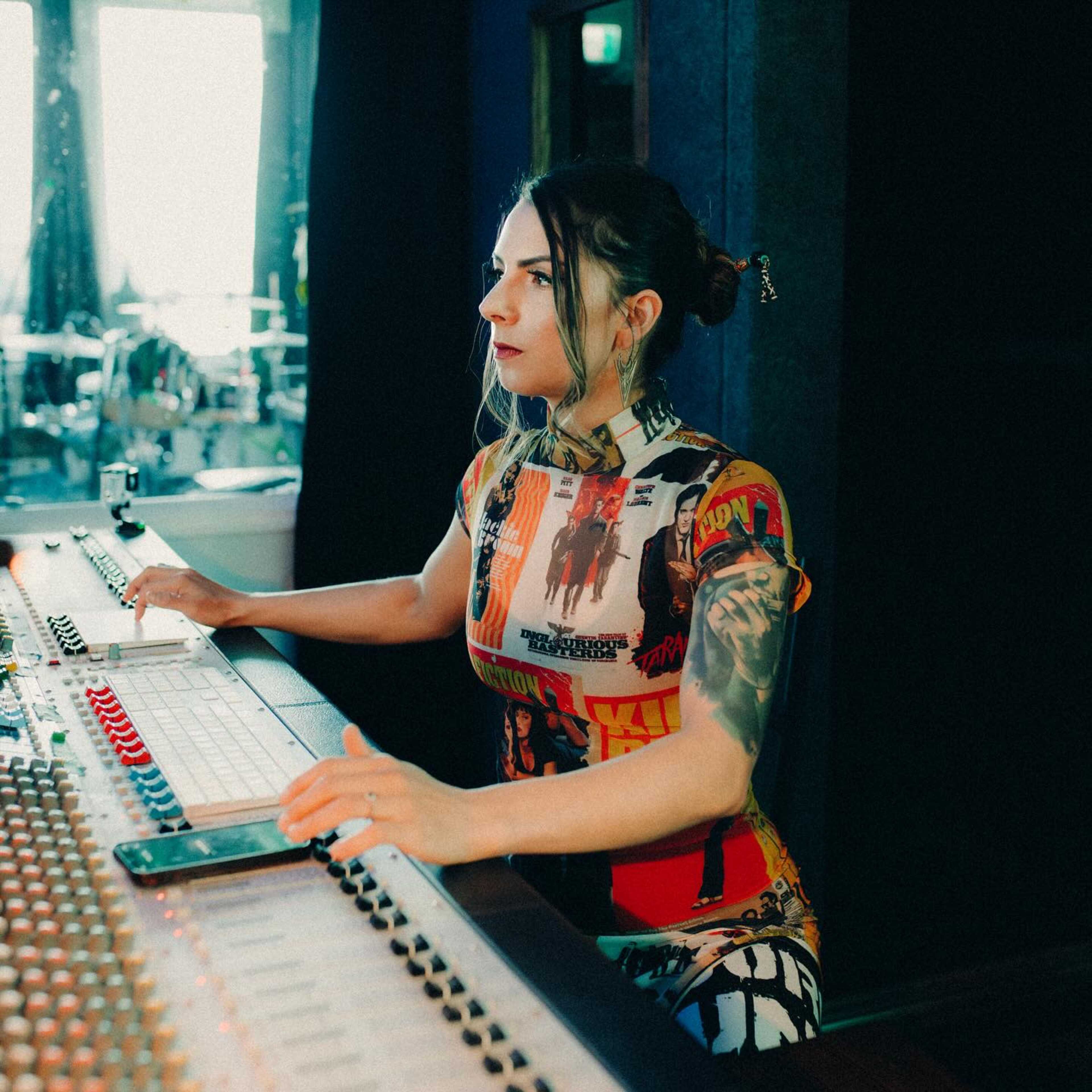 A woman with tattoos is operating a sound mixing console in a music studio.