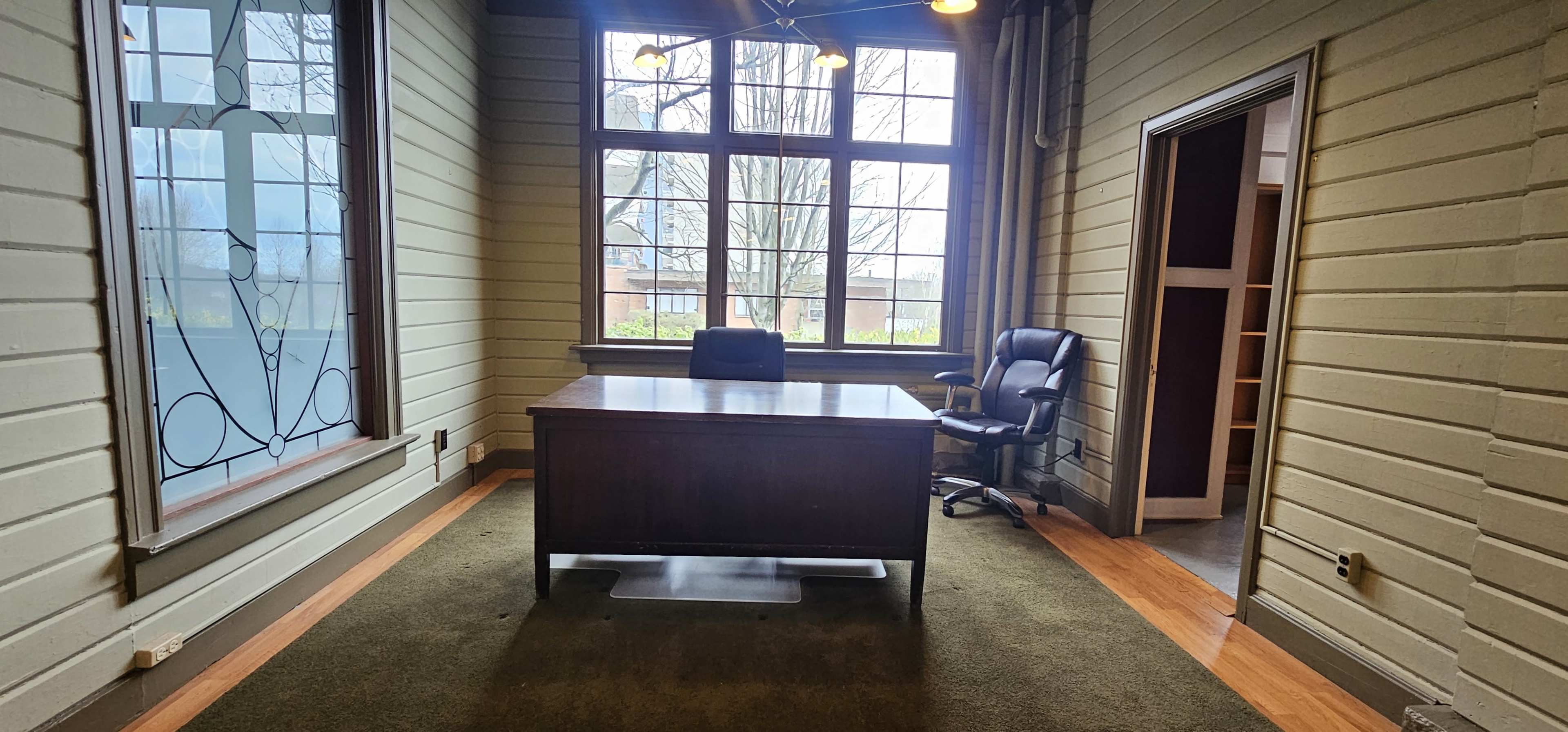 An empty office featuring a wooden desk and a black office chair, with large windows allowing natural light to enter.