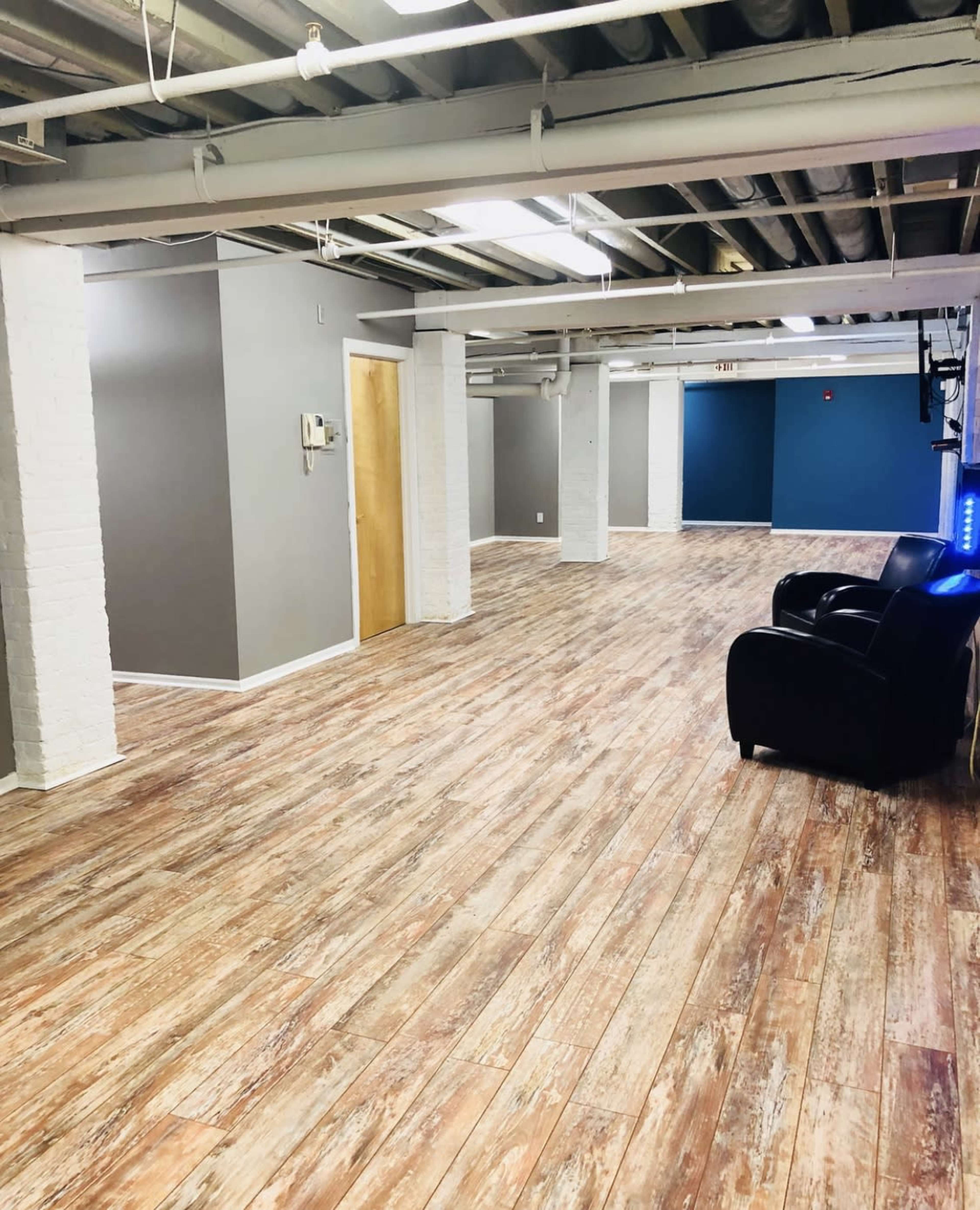 The image shows a spacious interior with a wooden floor, white brick columns, and gray walls, featuring a dark chair in one corner.