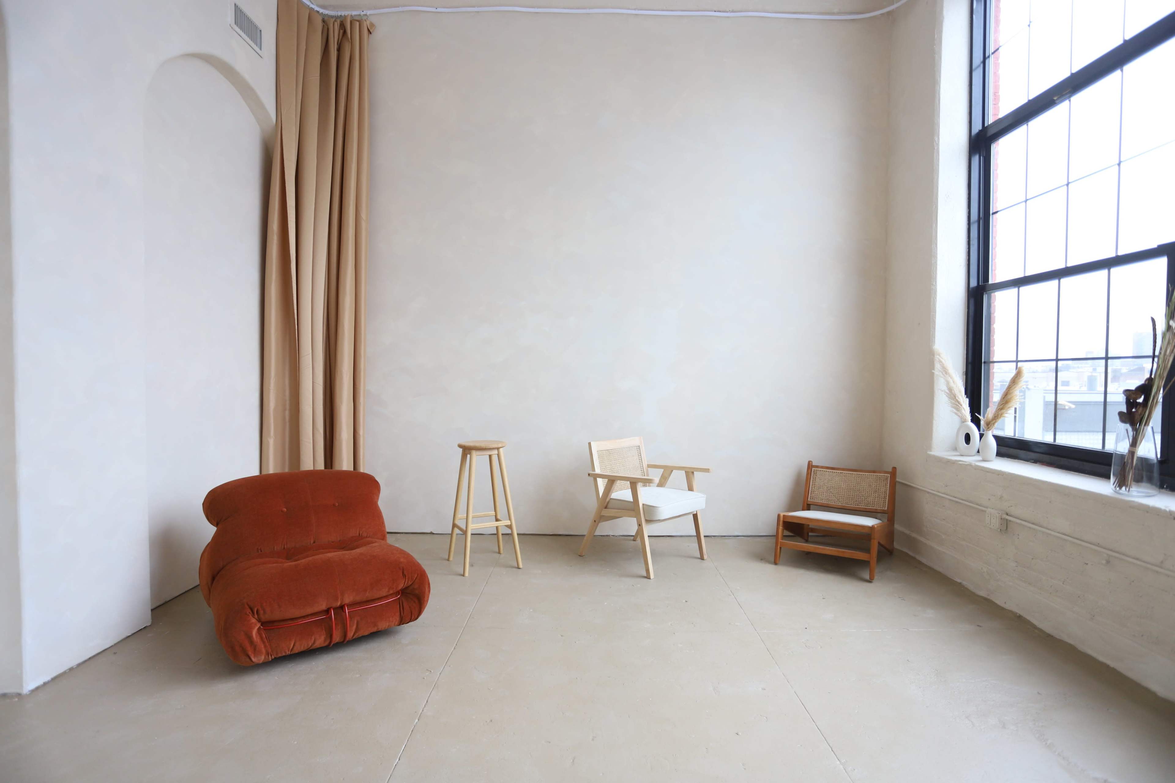 A minimalist interior space featuring an orange upholstered chair, a wooden stool, and two other various styled chairs near a large window.