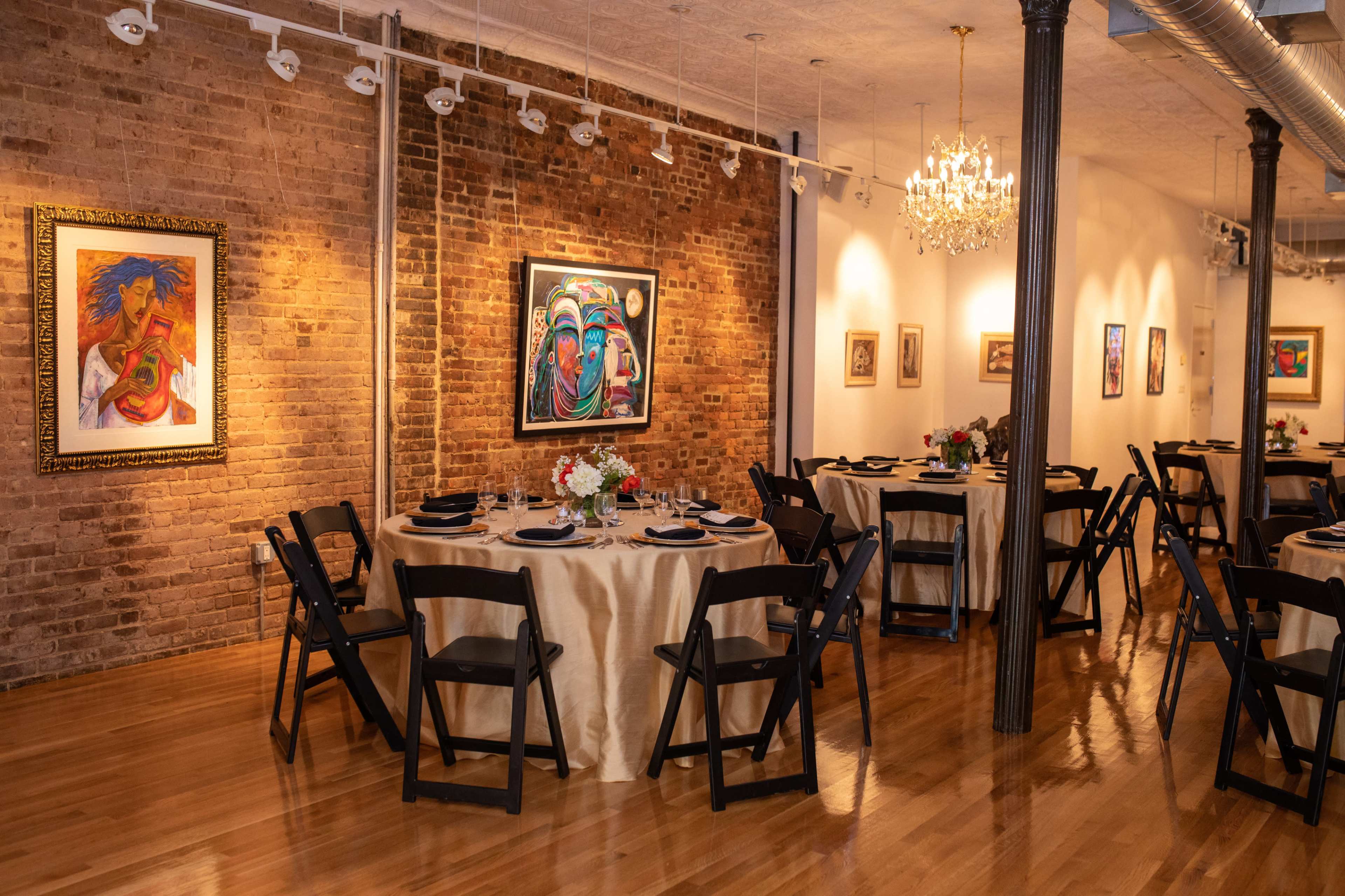 The image shows an elegant dining setup with round tables adorned with cream tablecloths and black chairs in an art gallery featuring colorful paintings on exposed brick walls.