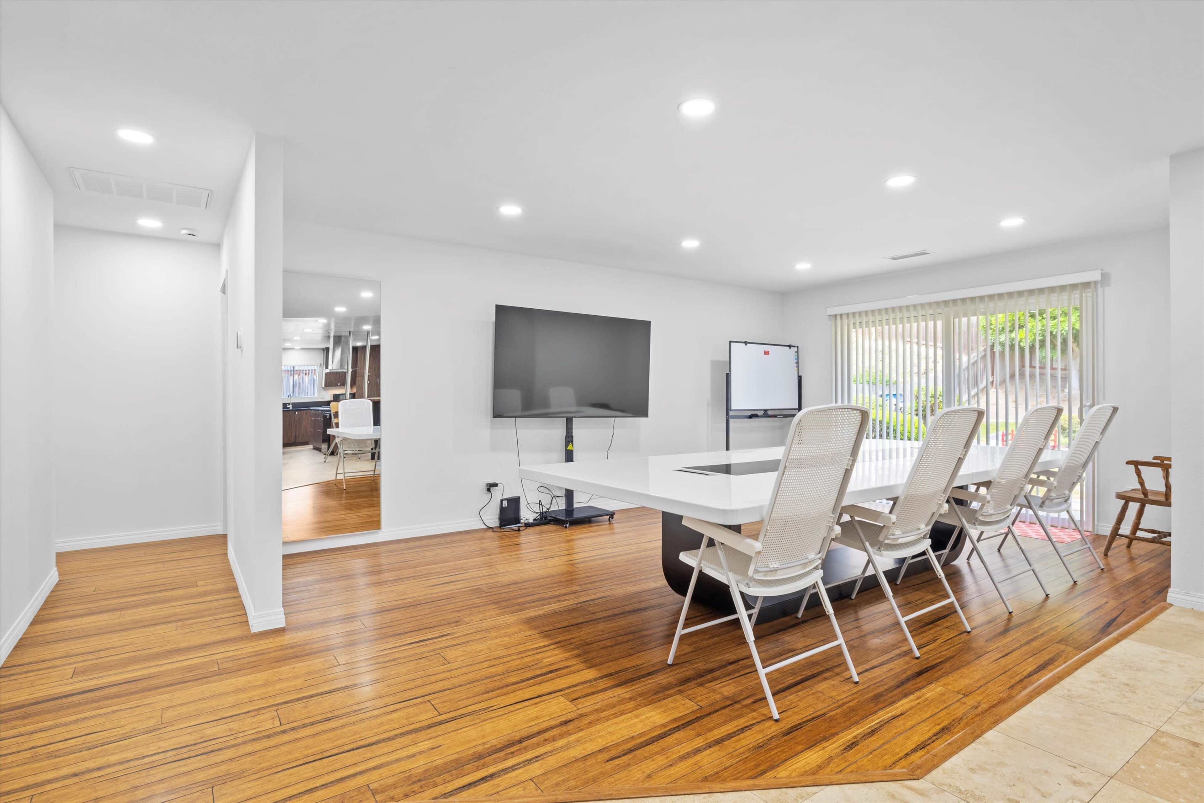 A modern meeting room features a large white table surrounded by six white chairs, with a flat-screen TV mounted on the wall and a sliding door leading to an outdoor space.