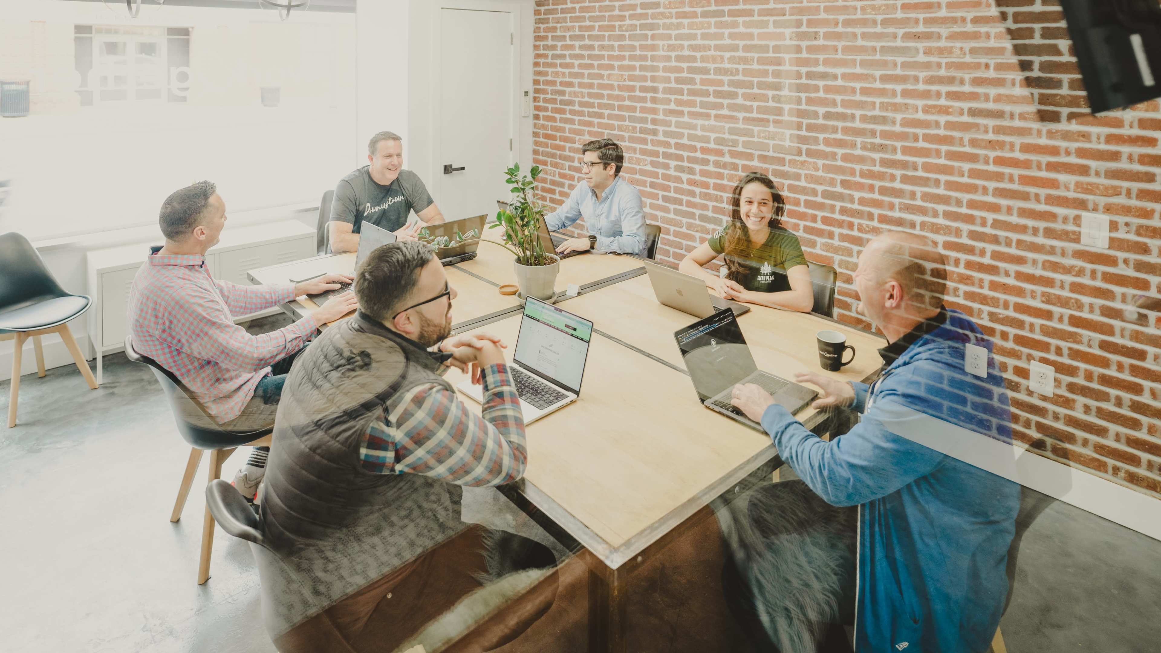 A group of six people is seated around a large wooden table in a meeting room with a brick wall, engaged in discussion and using laptops.