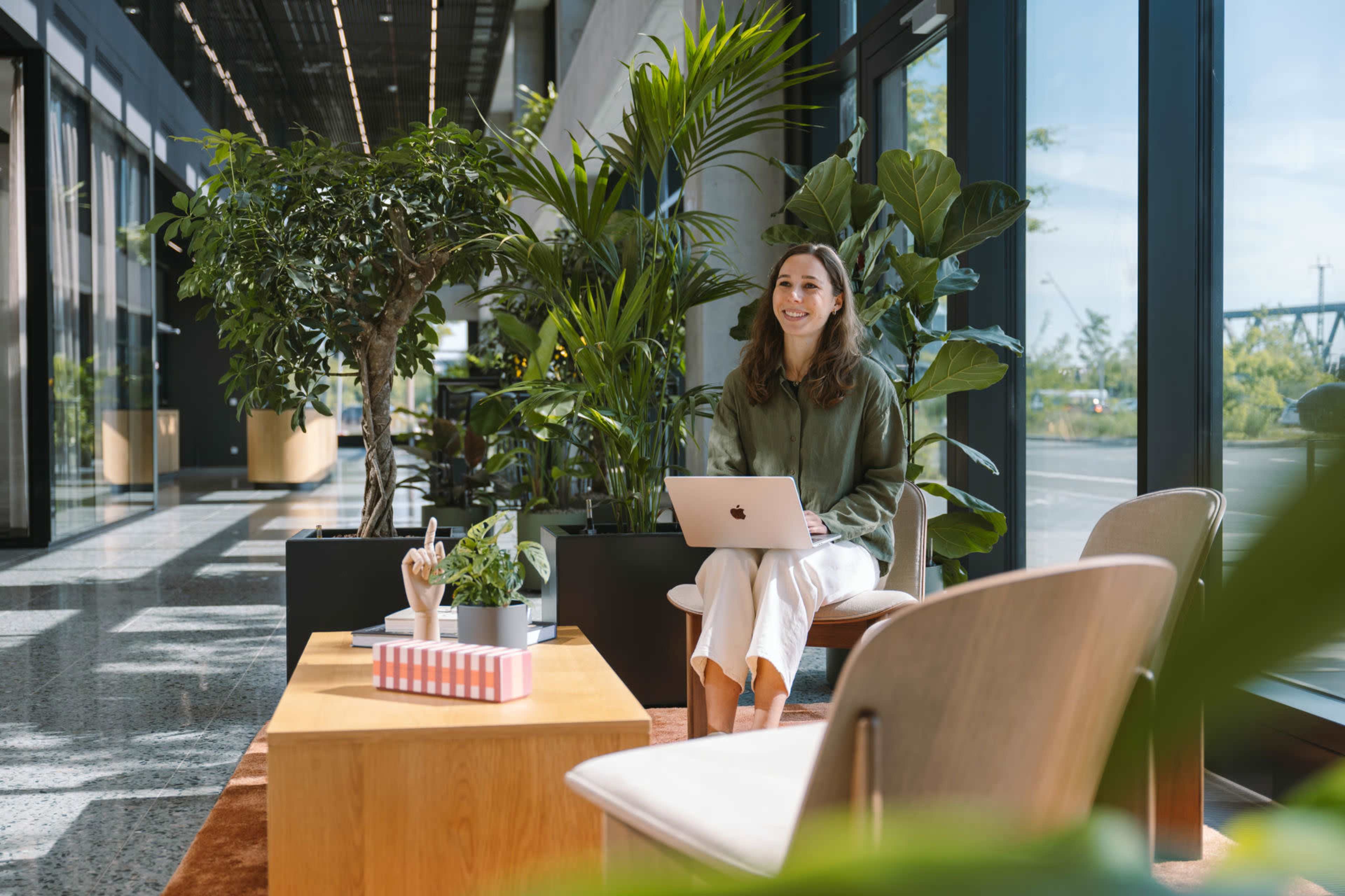 A woman sits on a bench with a laptop in a modern, plant-filled indoor space with large windows.