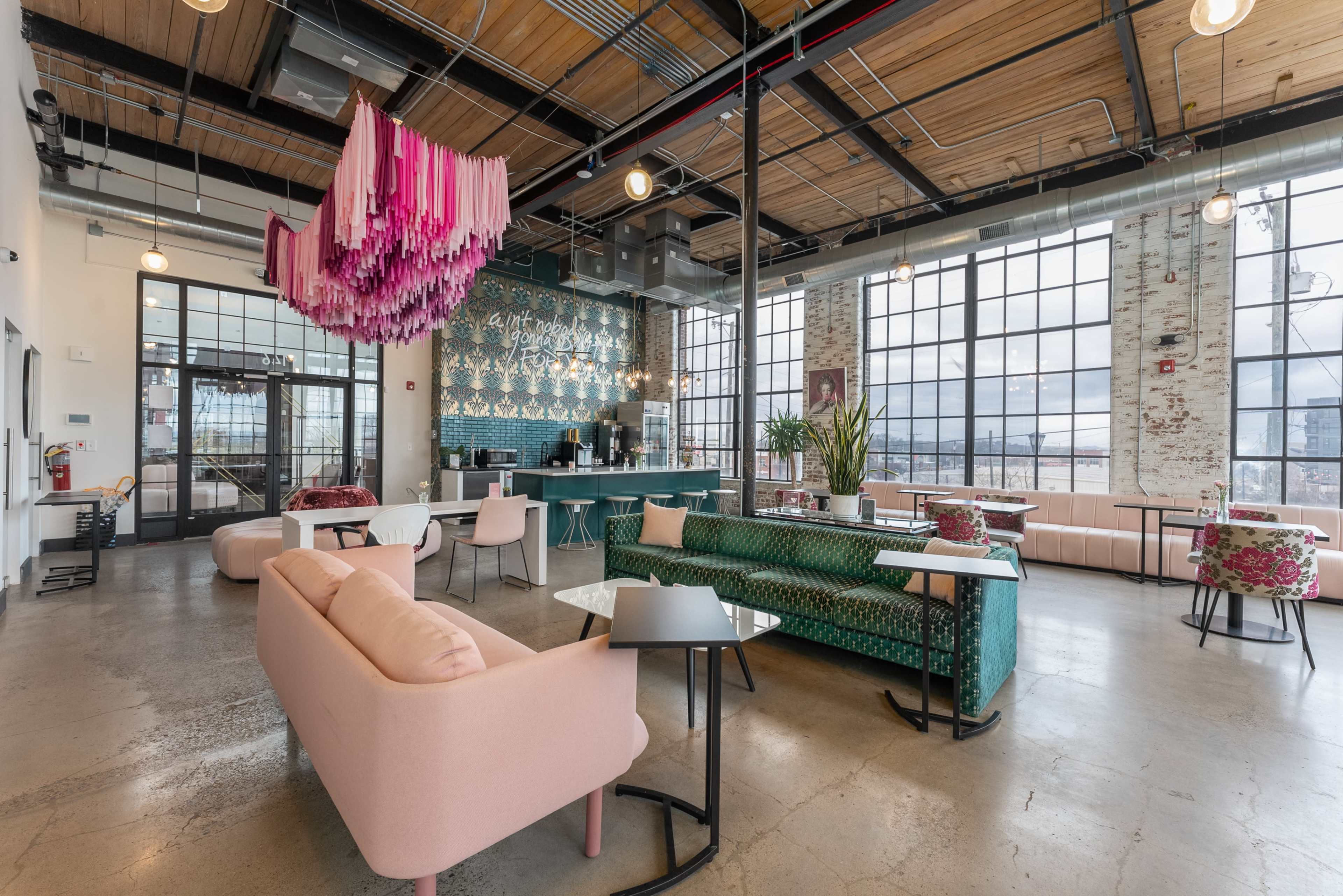 A modern social space with large windows, various seating areas, a vibrant ceiling installation, and a bar counter in the background.