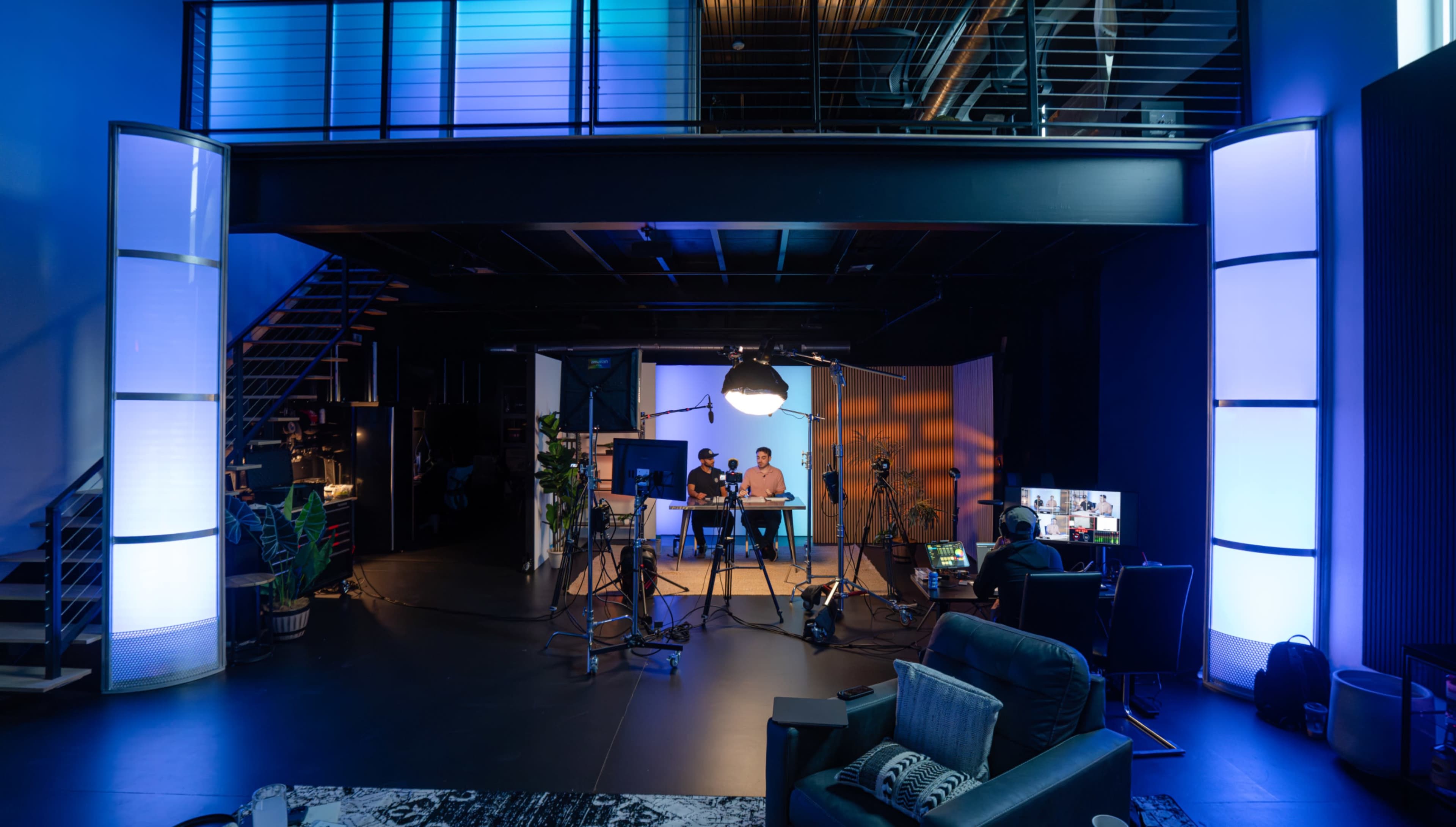 The image shows a modern studio space with multiple cameras and lighting equipment set up for a video production, featuring two people sitting at a desk in the center.