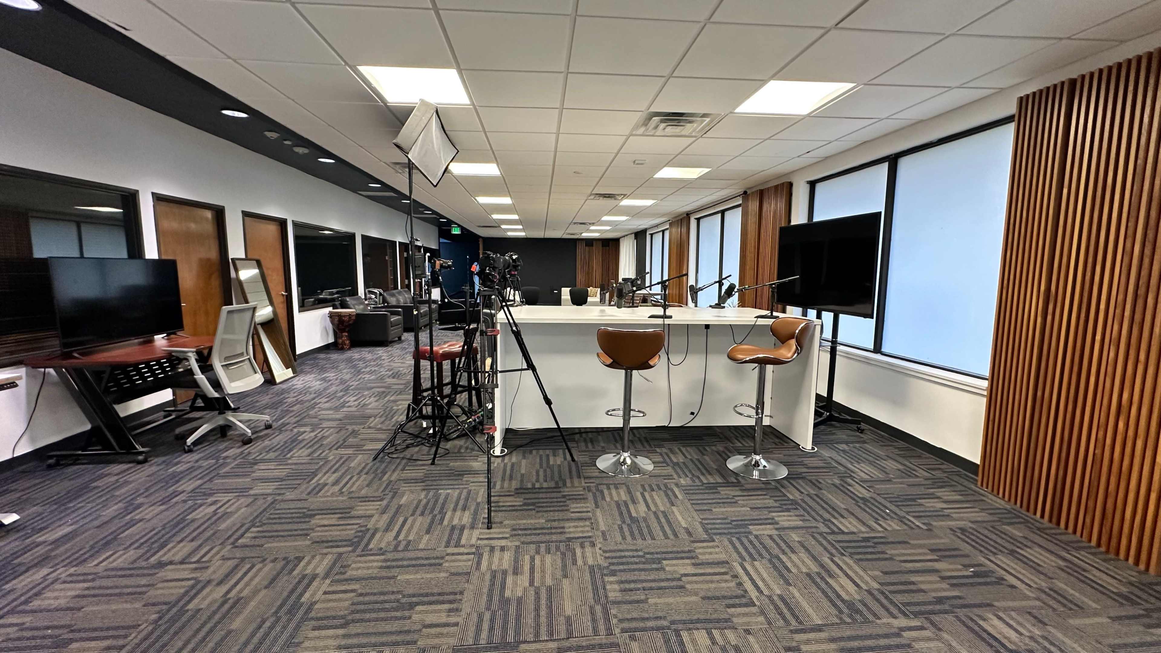 The image shows a modern office space with a bar-style counter, high stools, and equipment set up for video recording, accompanied by large windows and a contemporary design.