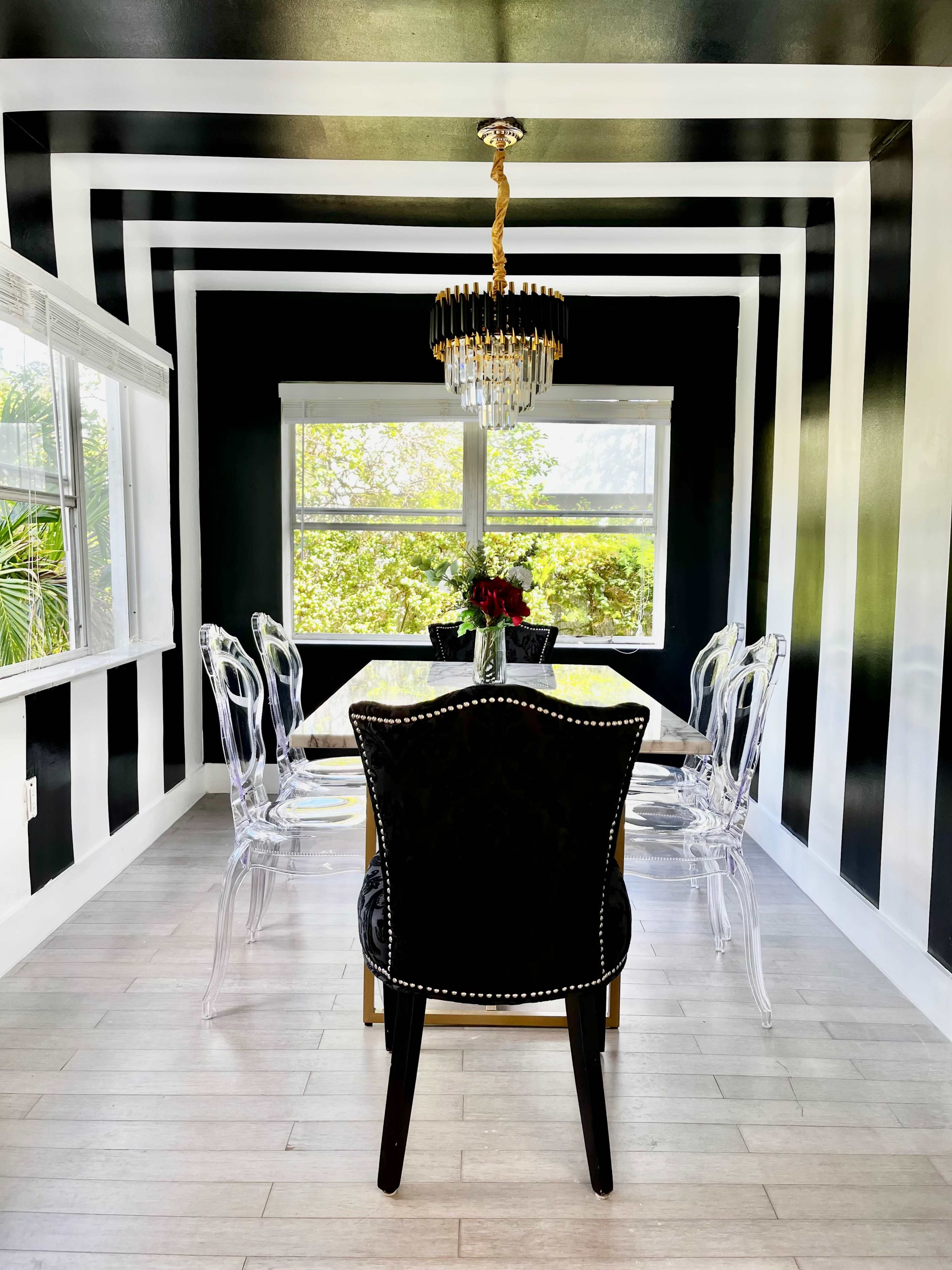 A dining room features a striped black and white wall design, a crystal chandelier, a glass table, and transparent chairs.