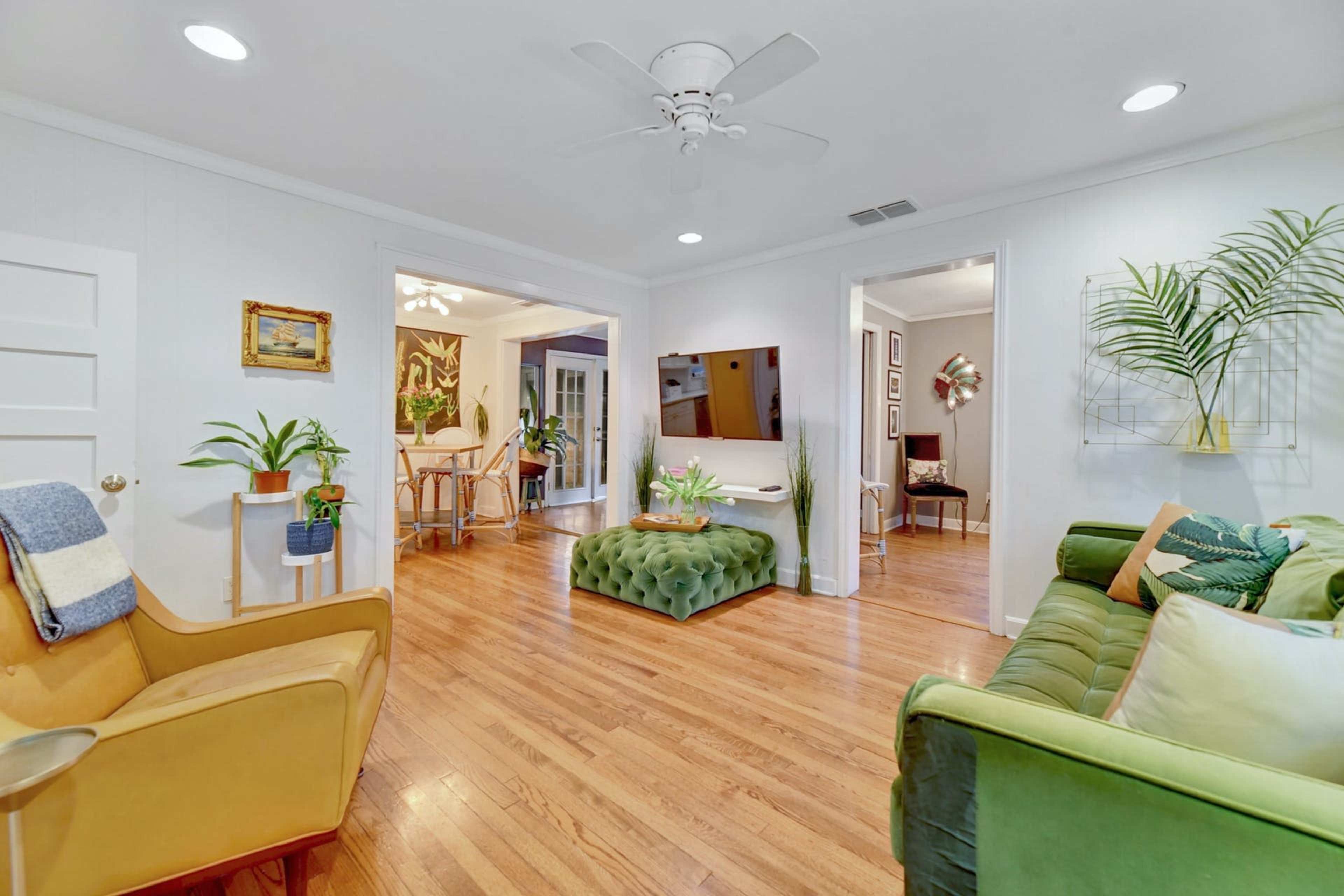 The image shows a bright living room featuring a green sofa, a tufted ottoman, and a doorway leading to another room with a dining area.