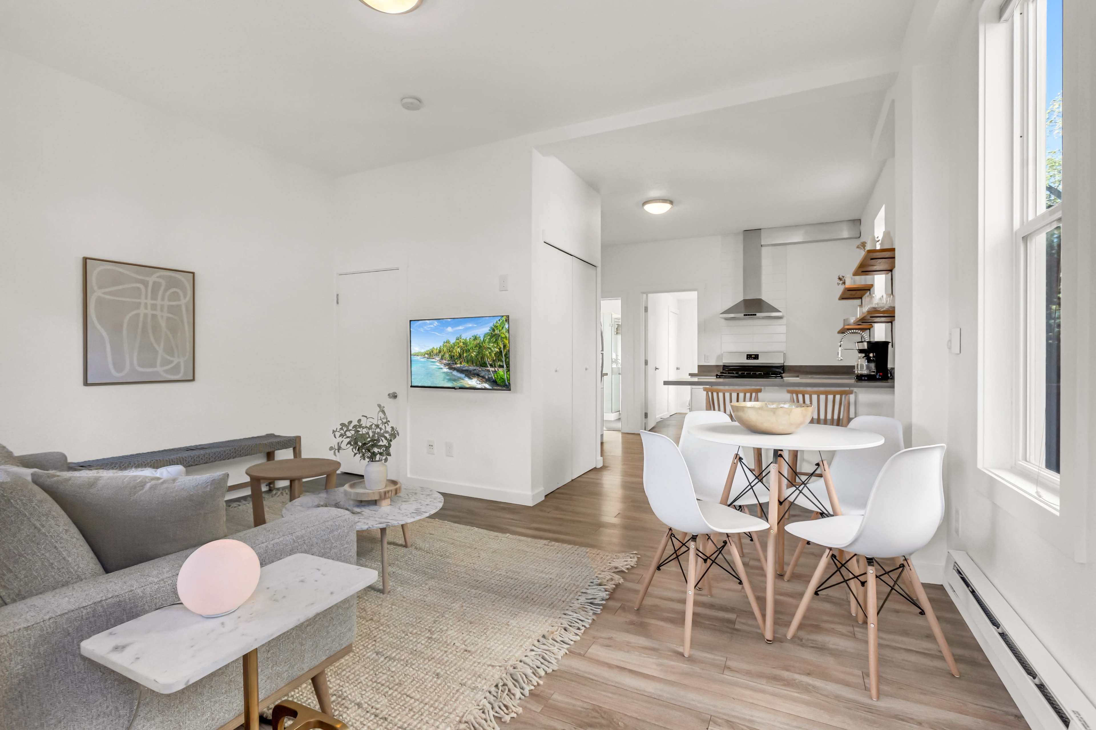 The image shows a modern living area with a gray sofa, a small round table with white chairs, and a wall-mounted television, leading to an adjacent kitchen.