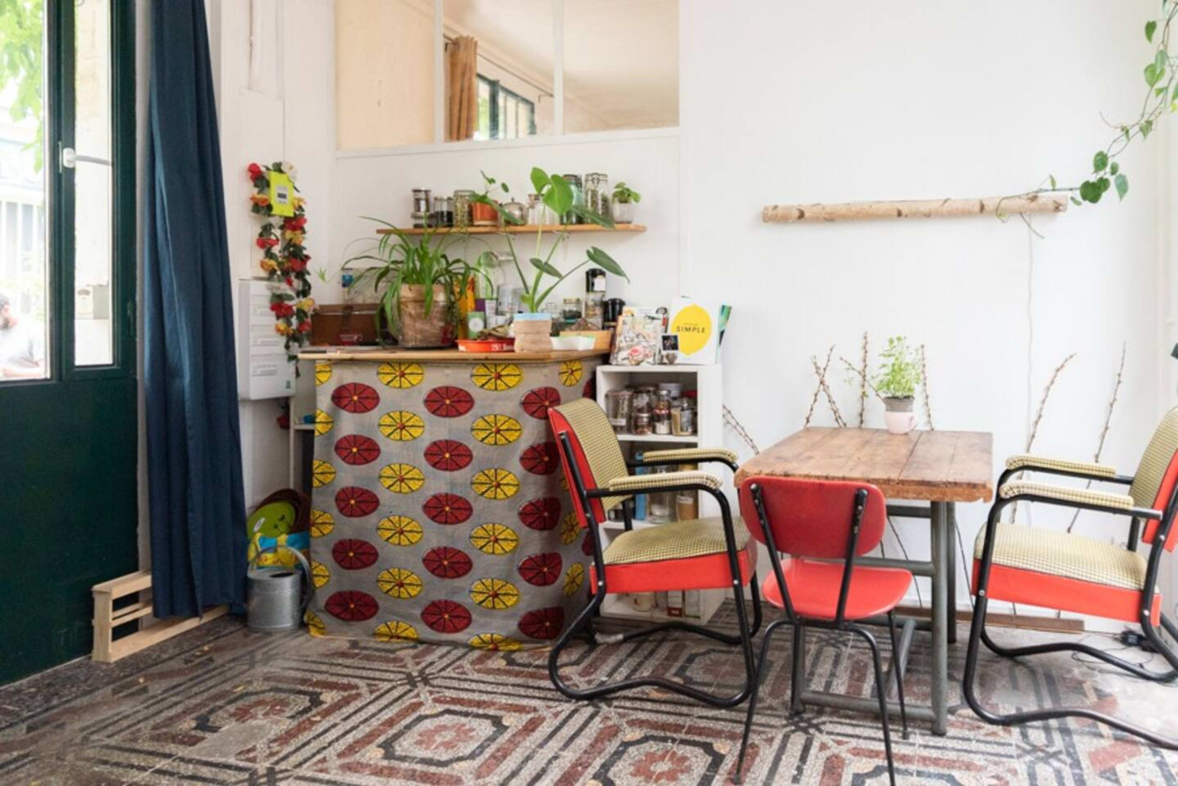 The image shows a cozy kitchen area with a patterned bar, a wooden table, and colorful chairs, surrounded by plants and decorative items.