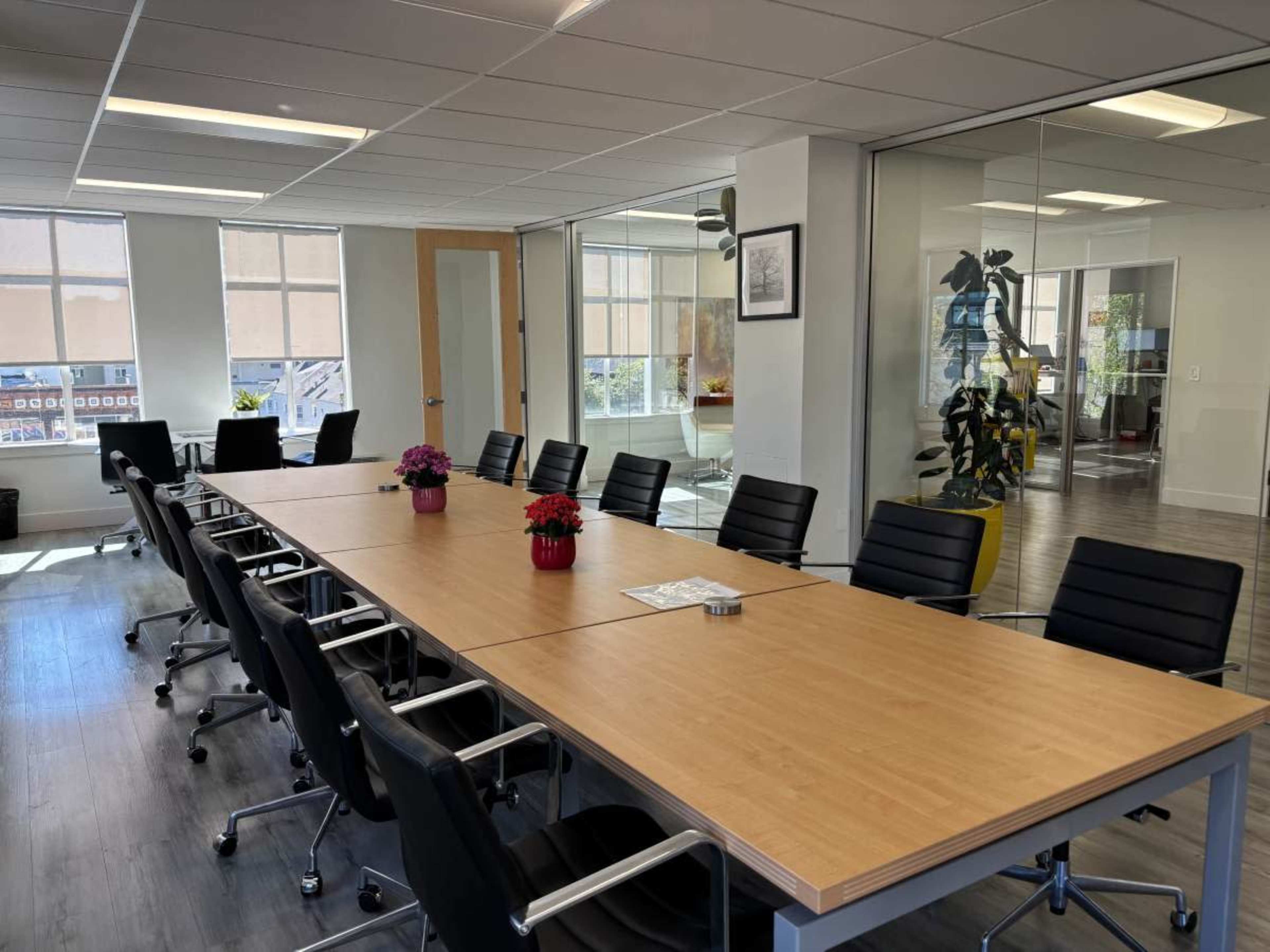 The image shows a modern conference room with a long wooden table surrounded by black chairs and large windows letting in natural light.