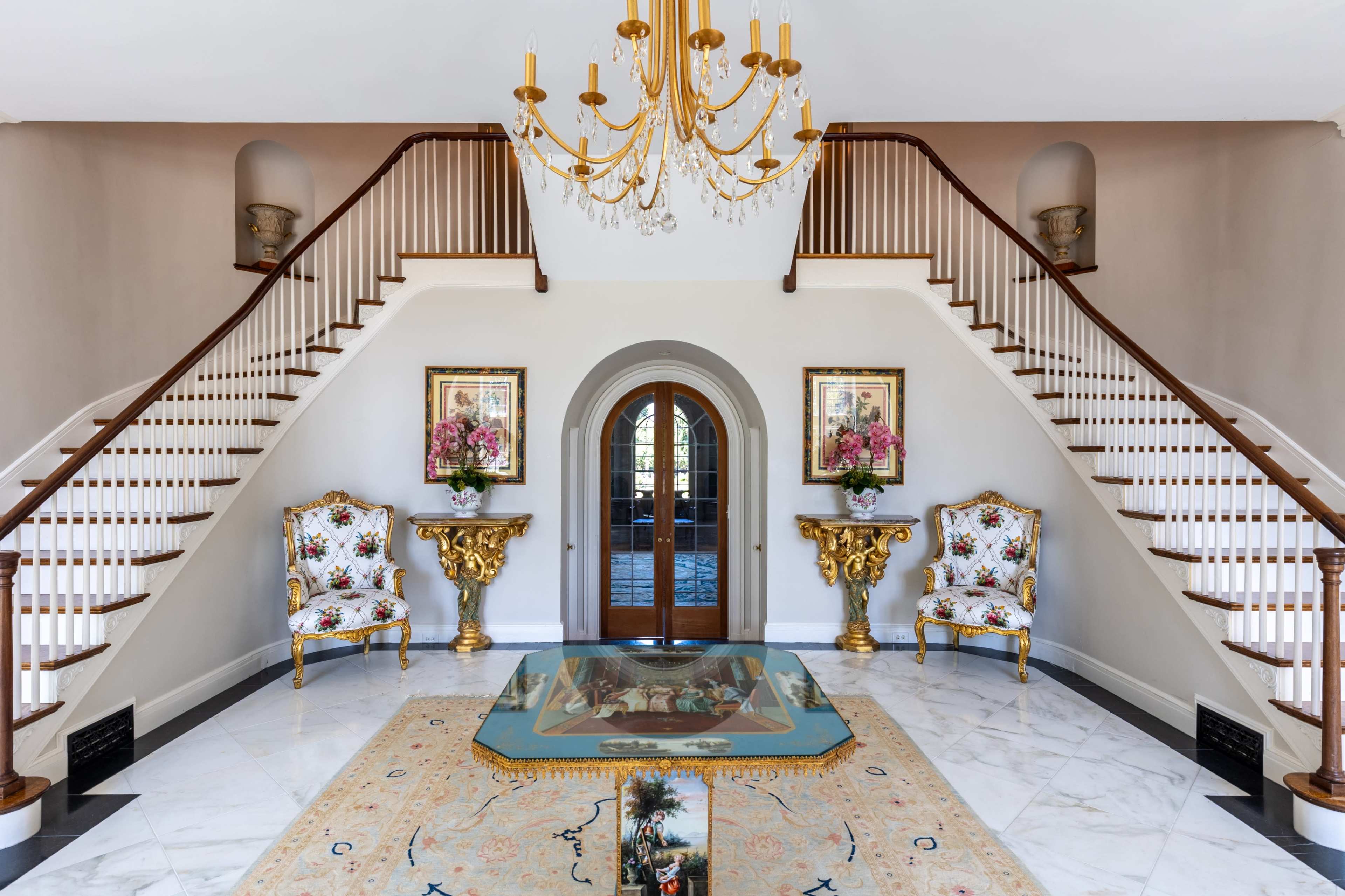 A grand entrance hall features a double staircase, elegant chandeliers, and ornate furniture framed by large windows.