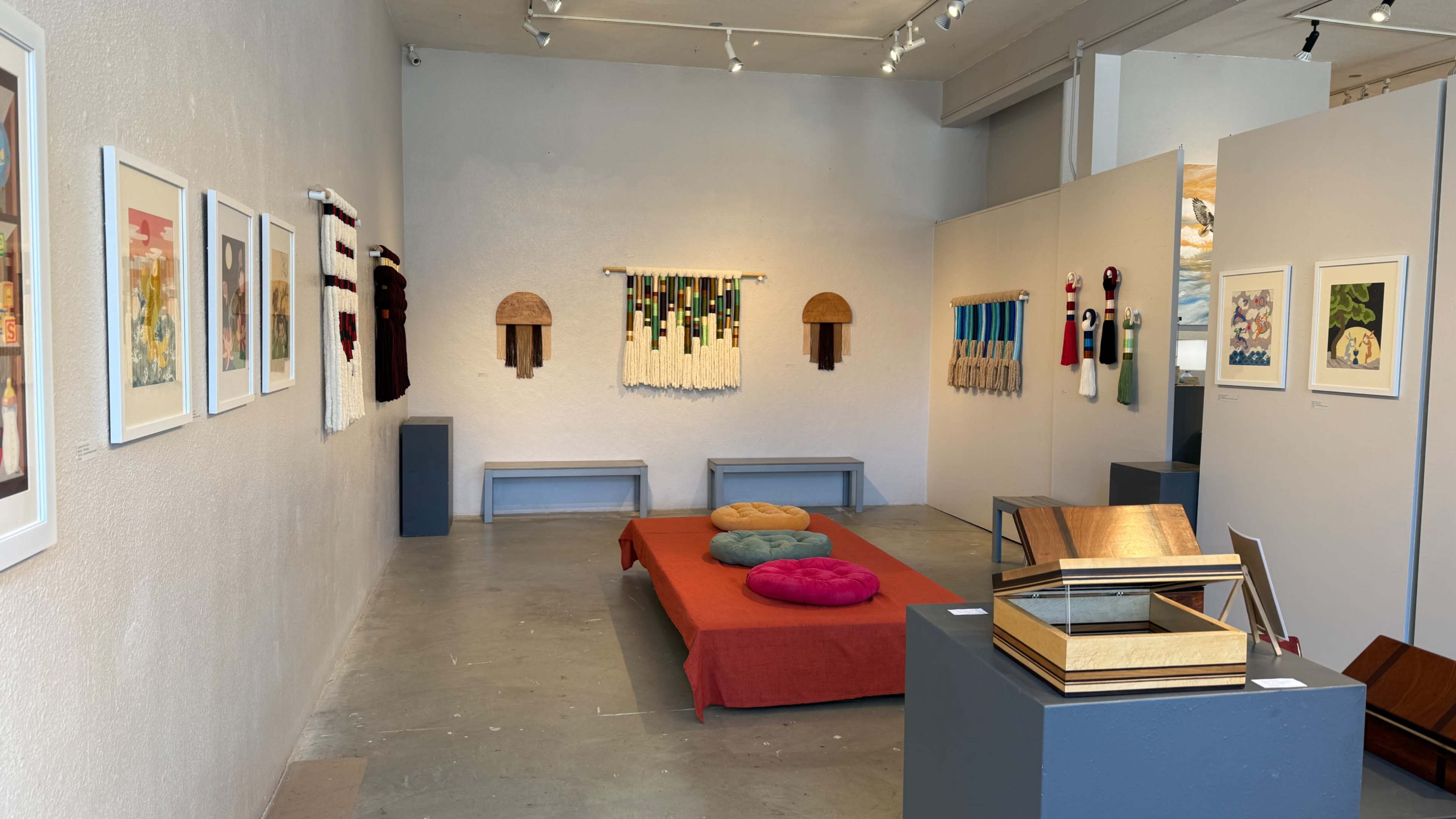 A spacious art gallery features colorful textile artworks on the walls, a red couch with cushions in the center, and a display stand on the right.