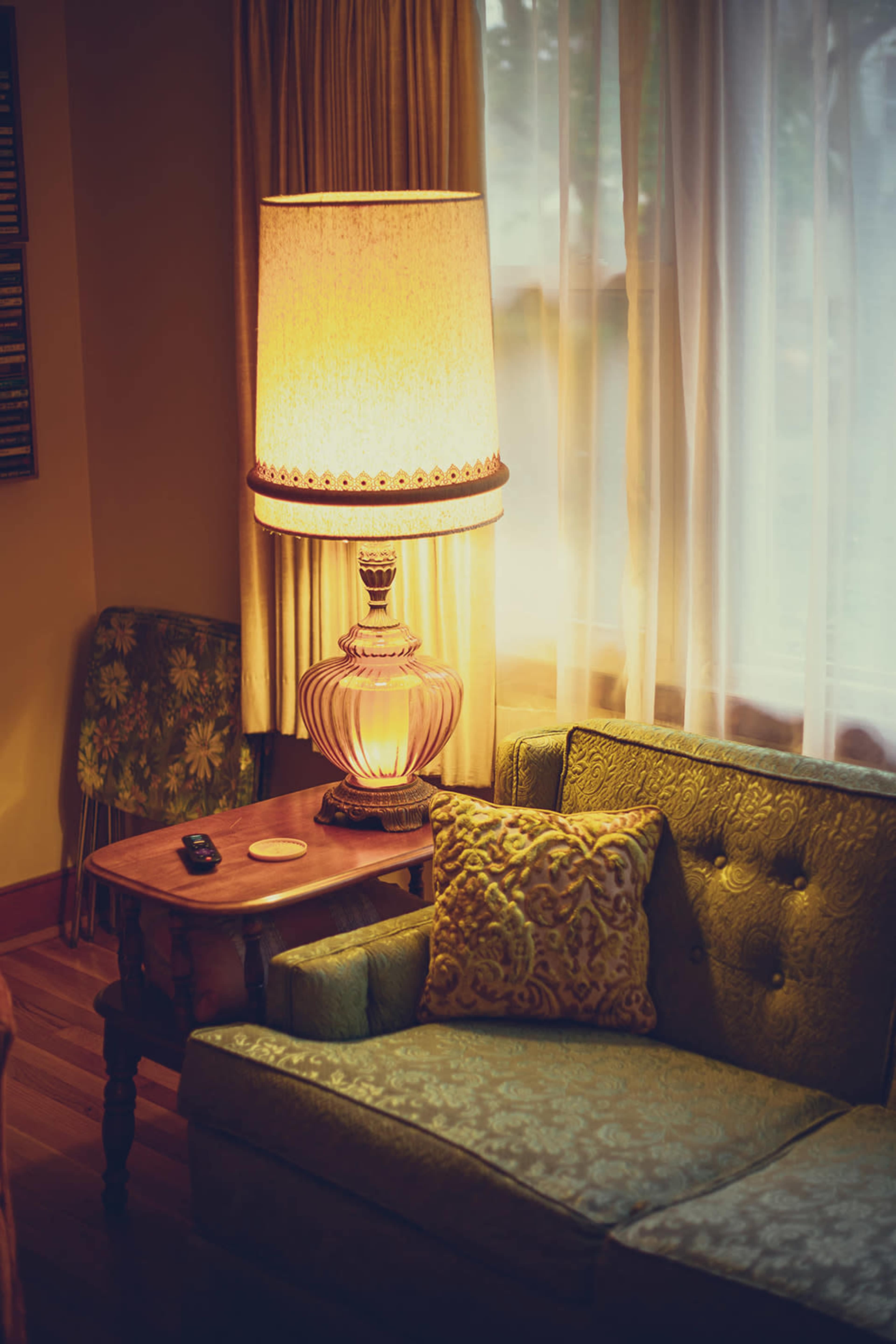A vintage living room features a green upholstered sofa, a small wooden table with a remote, and a large lamp casting warm light beside a window with sheer curtains.