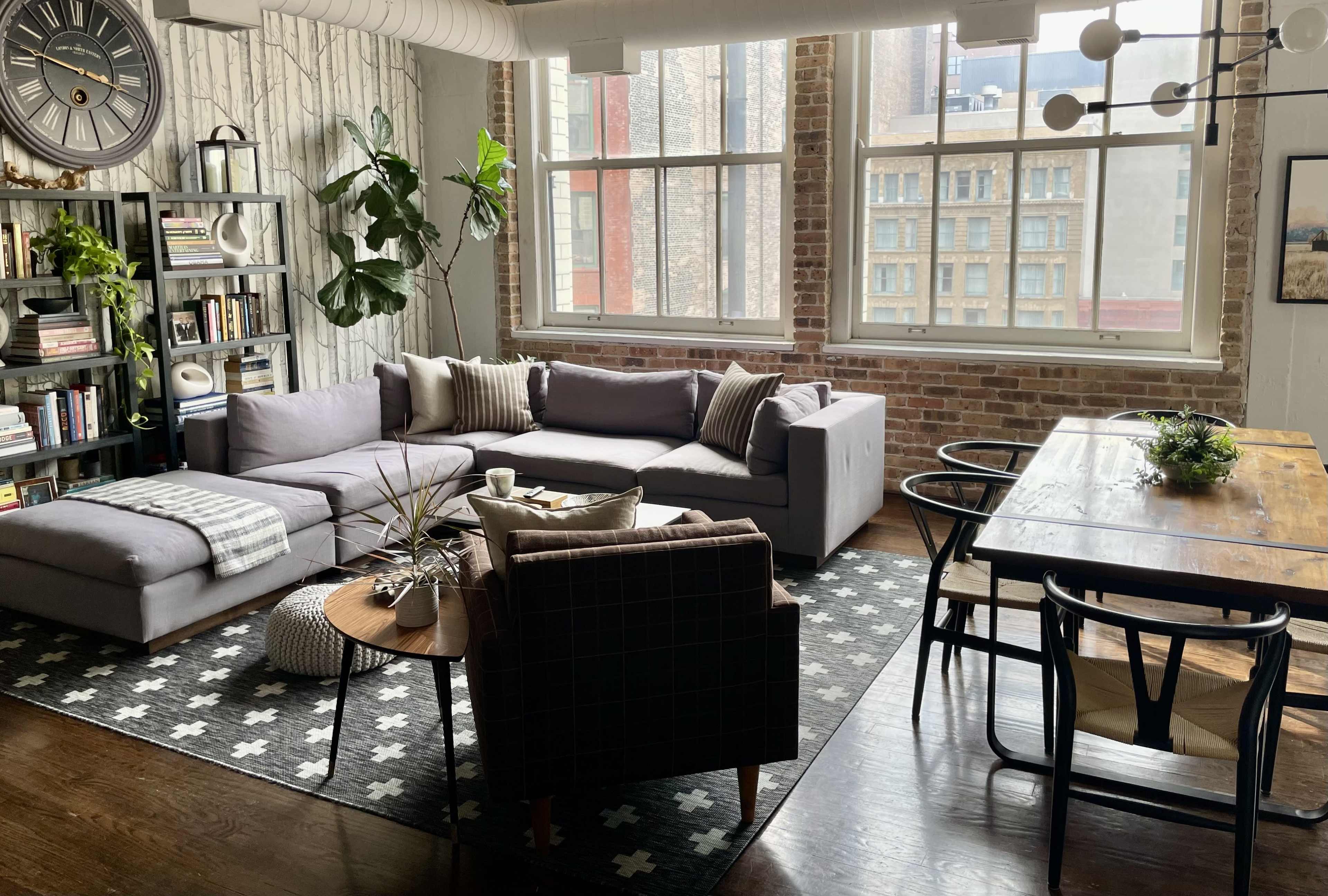 The image shows a cozy living space featuring a gray sectional sofa, a wooden dining table, bookshelves filled with books, and large windows that provide natural light.