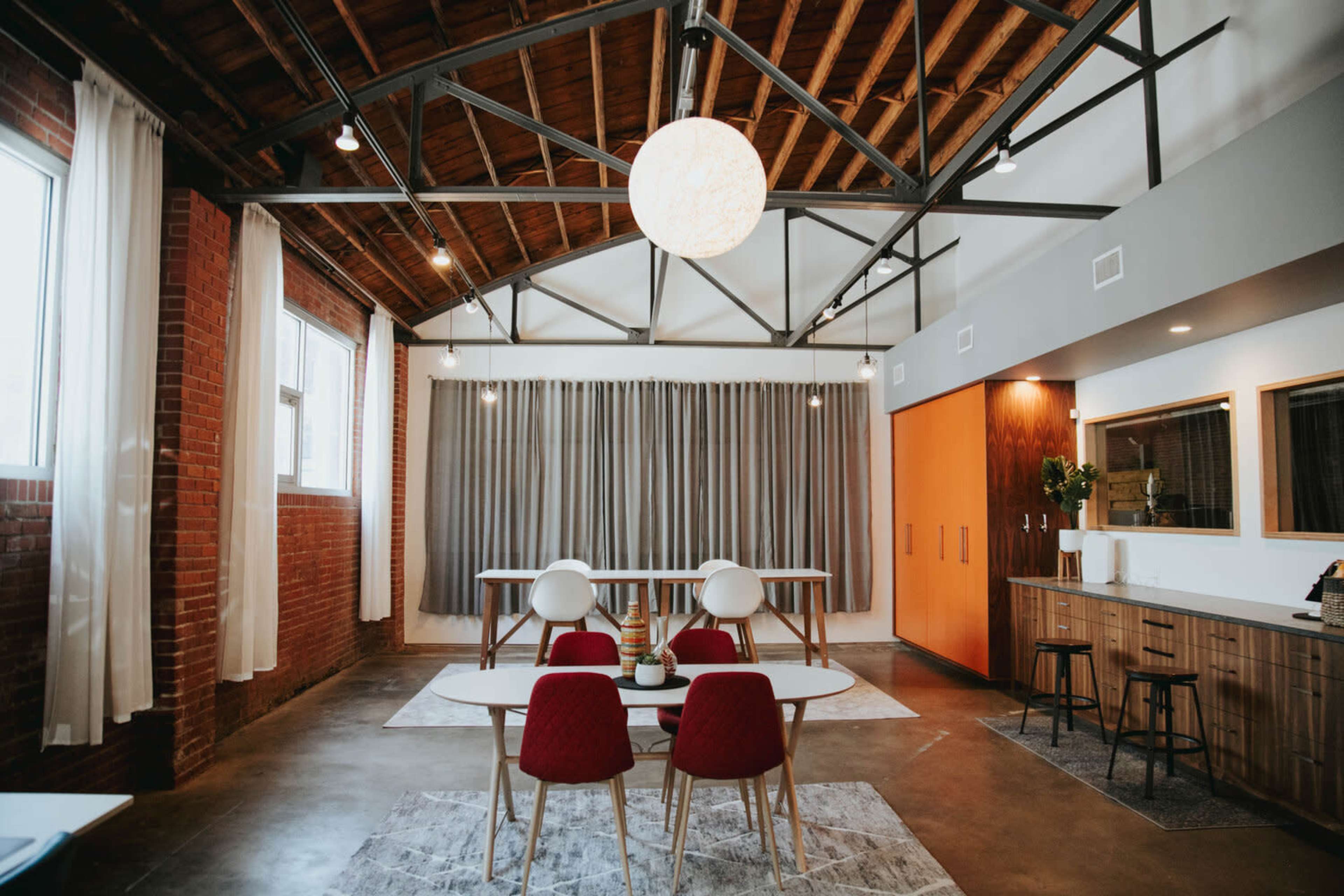 A modern, industrial-style room featuring a dining table with red chairs, a large round light fixture, and gray curtains in a space with exposed brick walls and wooden beams.