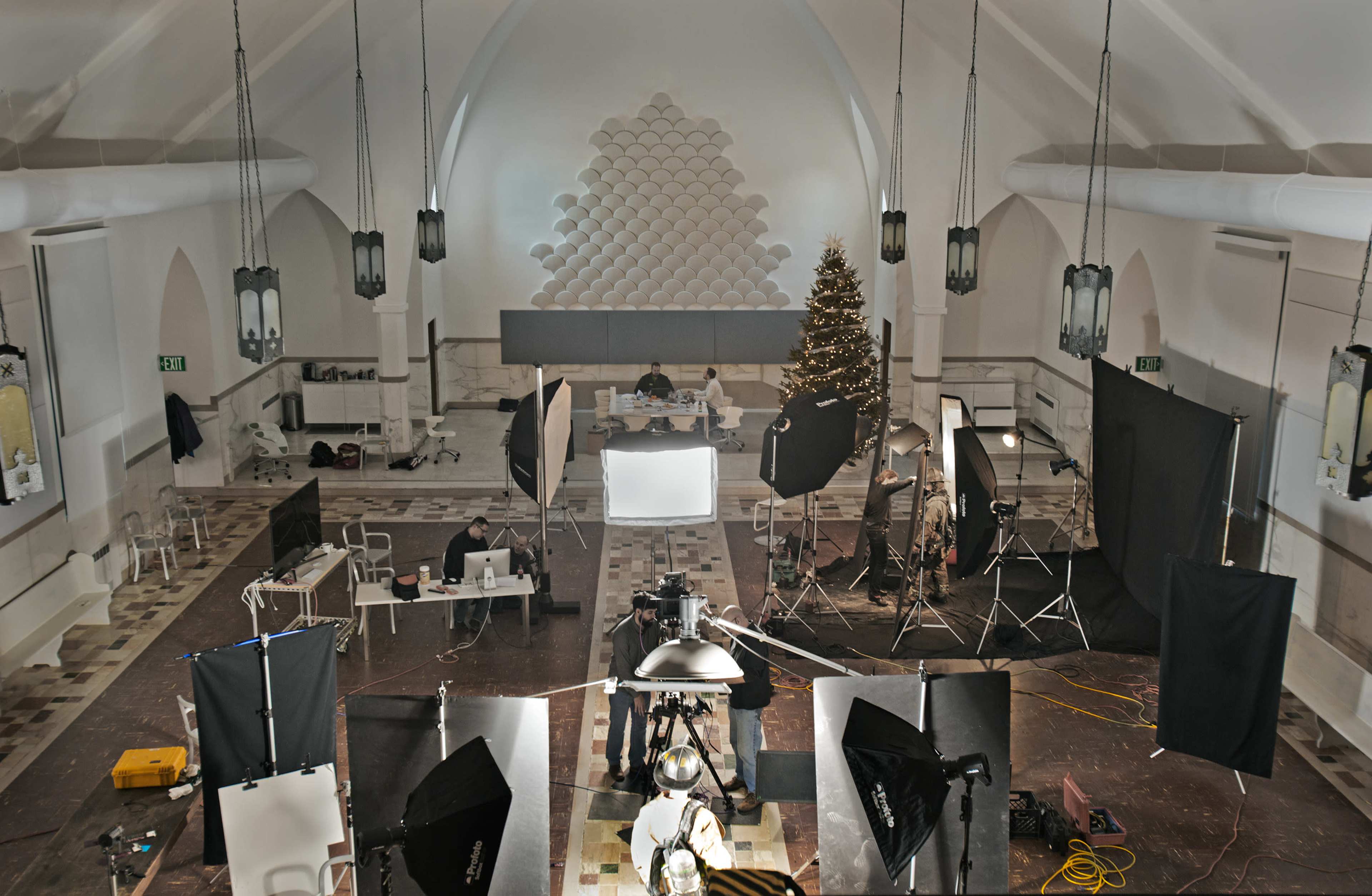 A large, well-lit studio space features multiple photography lights and equipment arranged around a central table with individuals working at computers, while a Christmas tree stands in the corner.