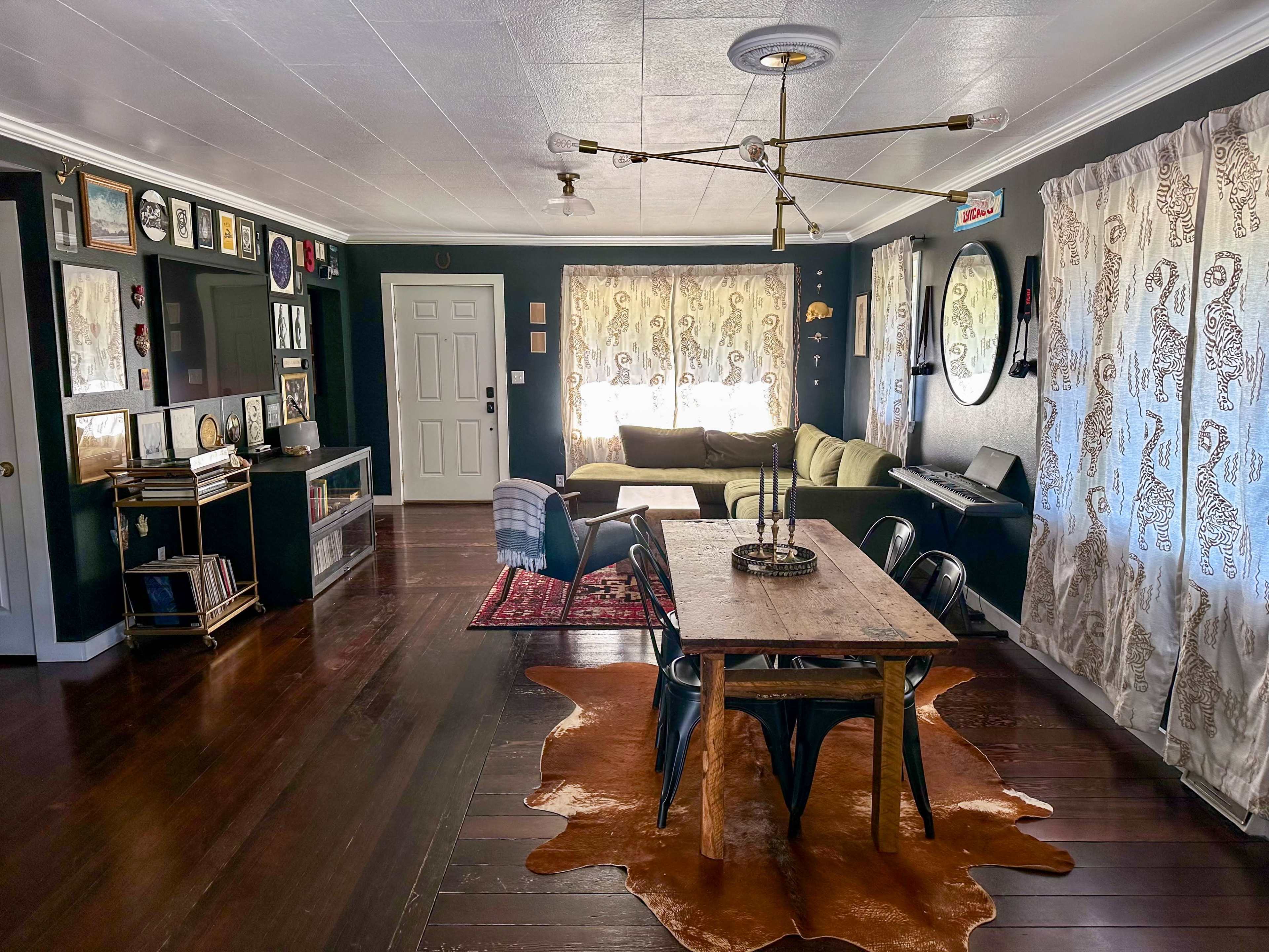 A living space with a dining area featuring a wooden table, black chairs, and a couch, adorned with patterned curtains and wooden flooring.