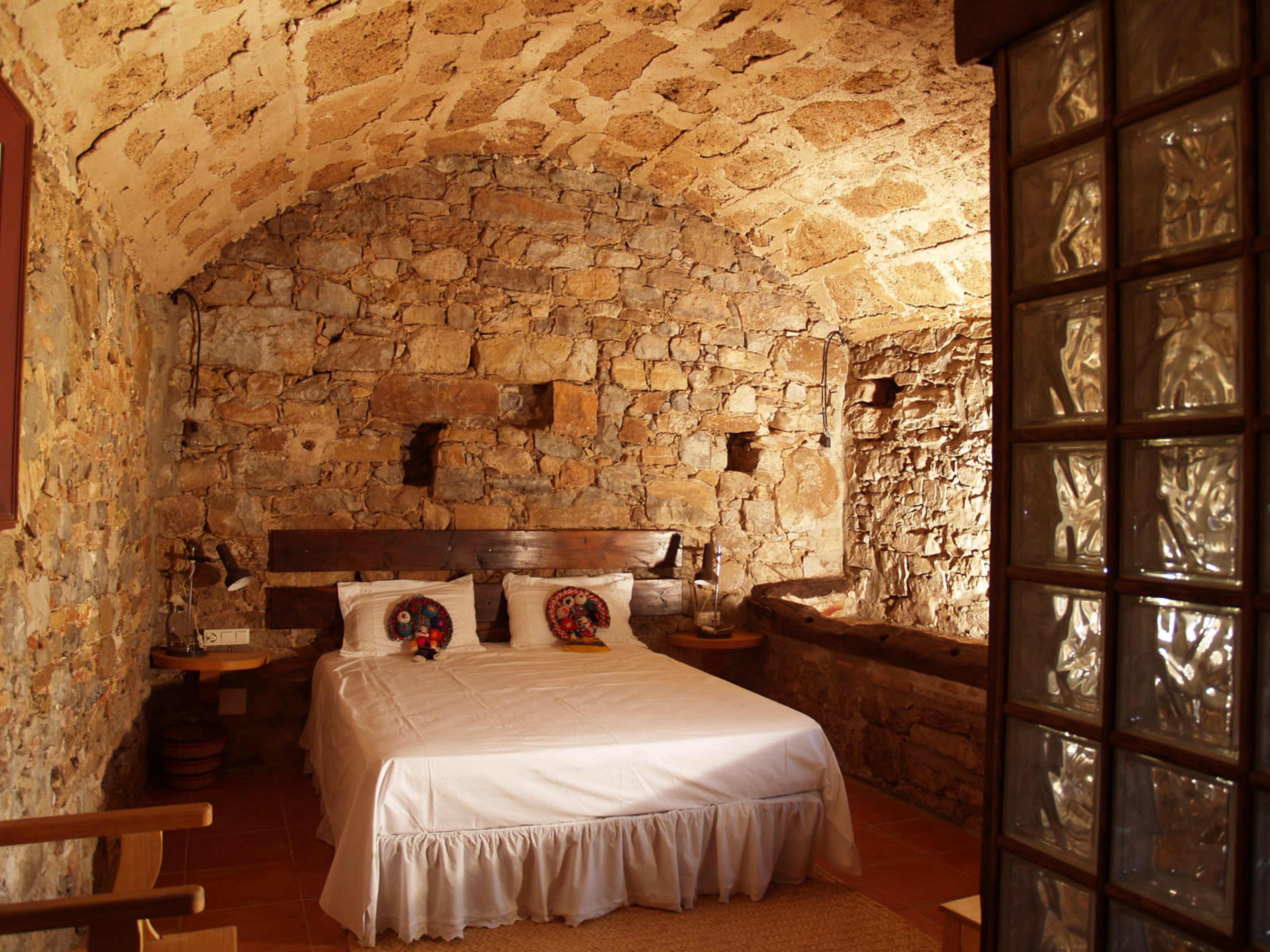The image shows a rustic bedroom featuring a bed with white linens against a stone wall, complemented by wooden furniture and a cozy atmosphere.