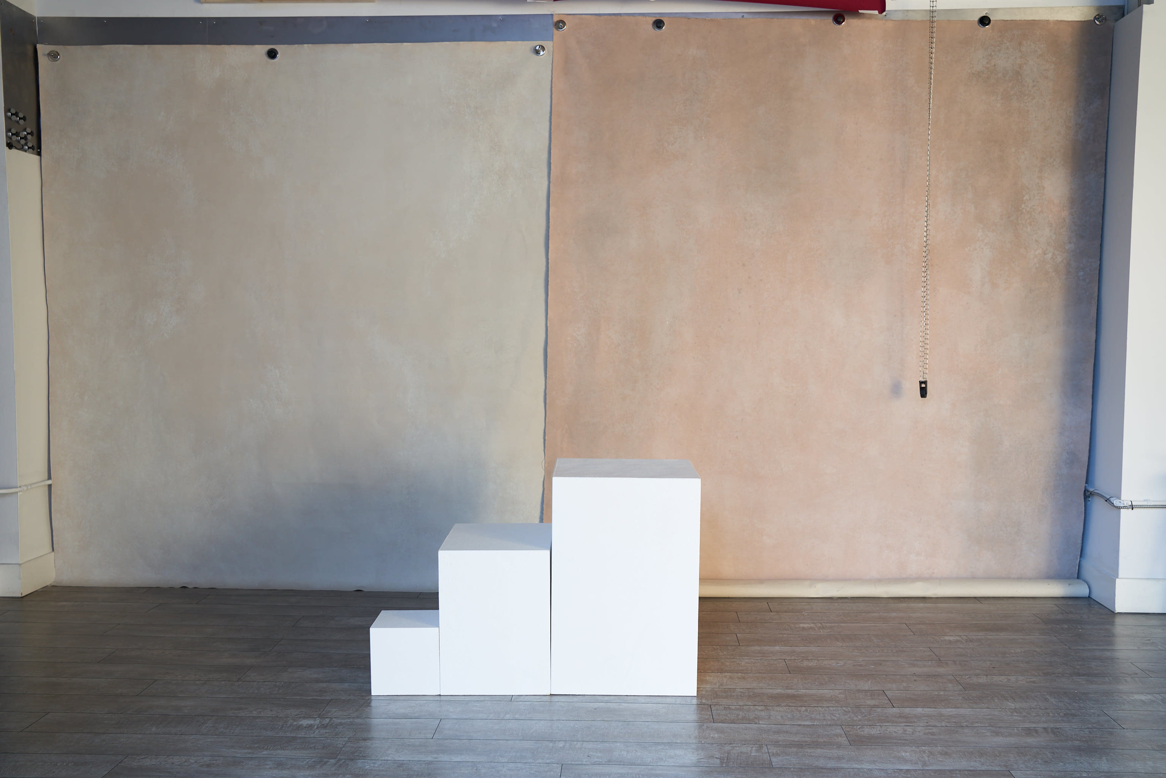 The image shows a minimal studio space featuring two textured backdrops—one gray and one pink—along with a set of three white cubes arranged in a stair-step formation on a wooden floor.