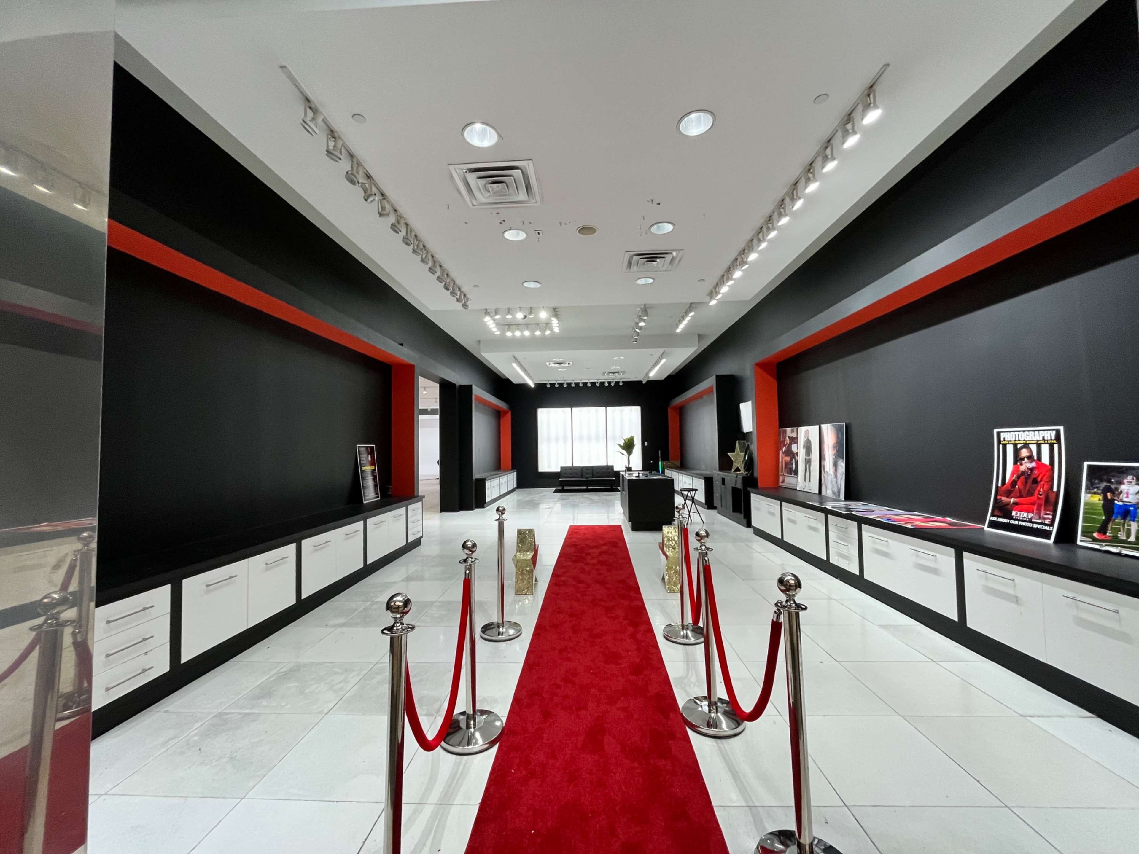 The image shows a modern exhibition space with a red carpet leading through a corridor flanked by black walls and white cabinets, featuring several framed photographs on display.