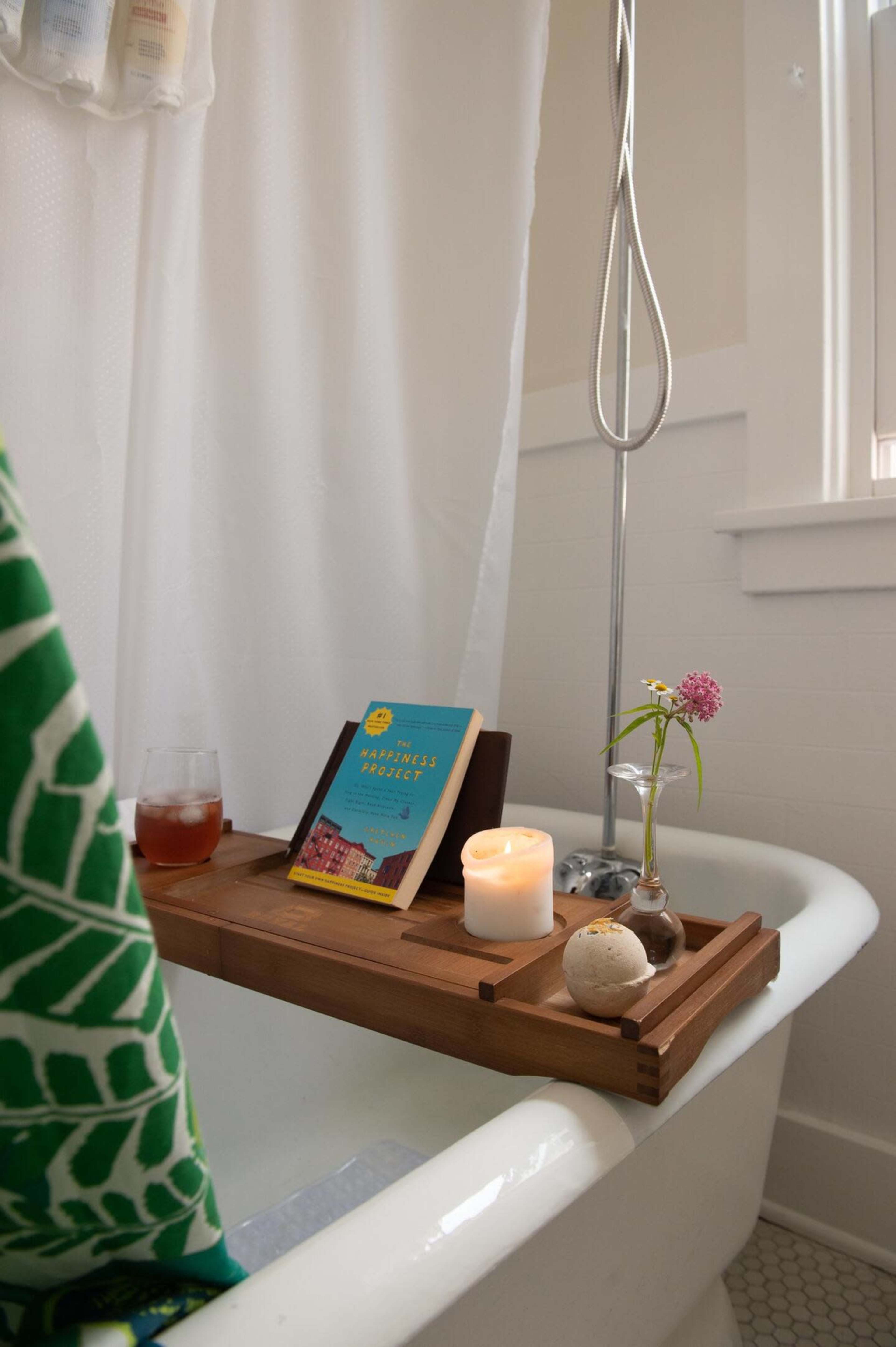A wooden bath tray holding a book, a candle, a glass of drink, and a small flower vase is placed across the edge of a clawfoot bathtub.