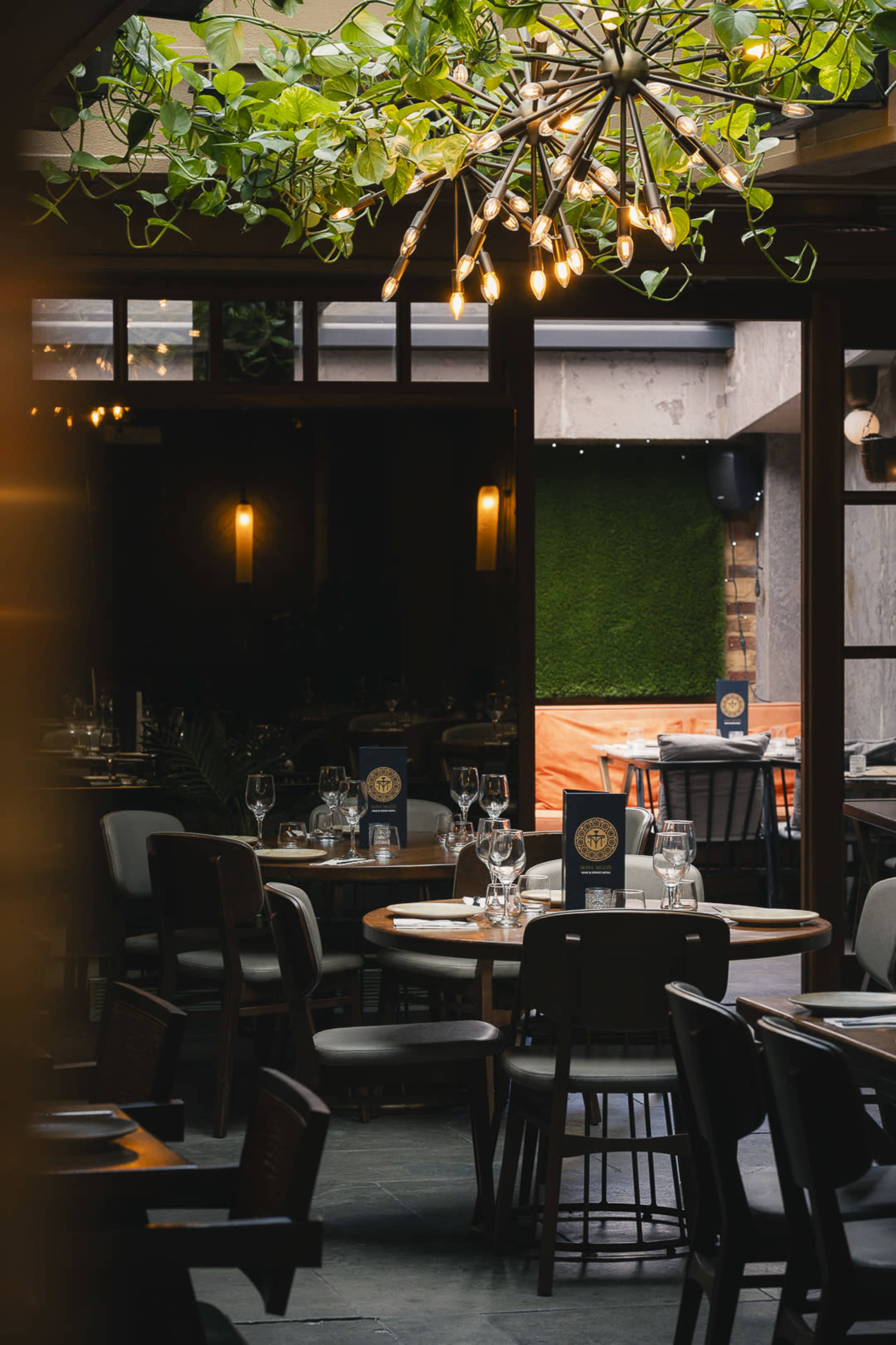 A restaurant interior features circular wooden tables, wine glasses, and a modern light fixture with greenery above.