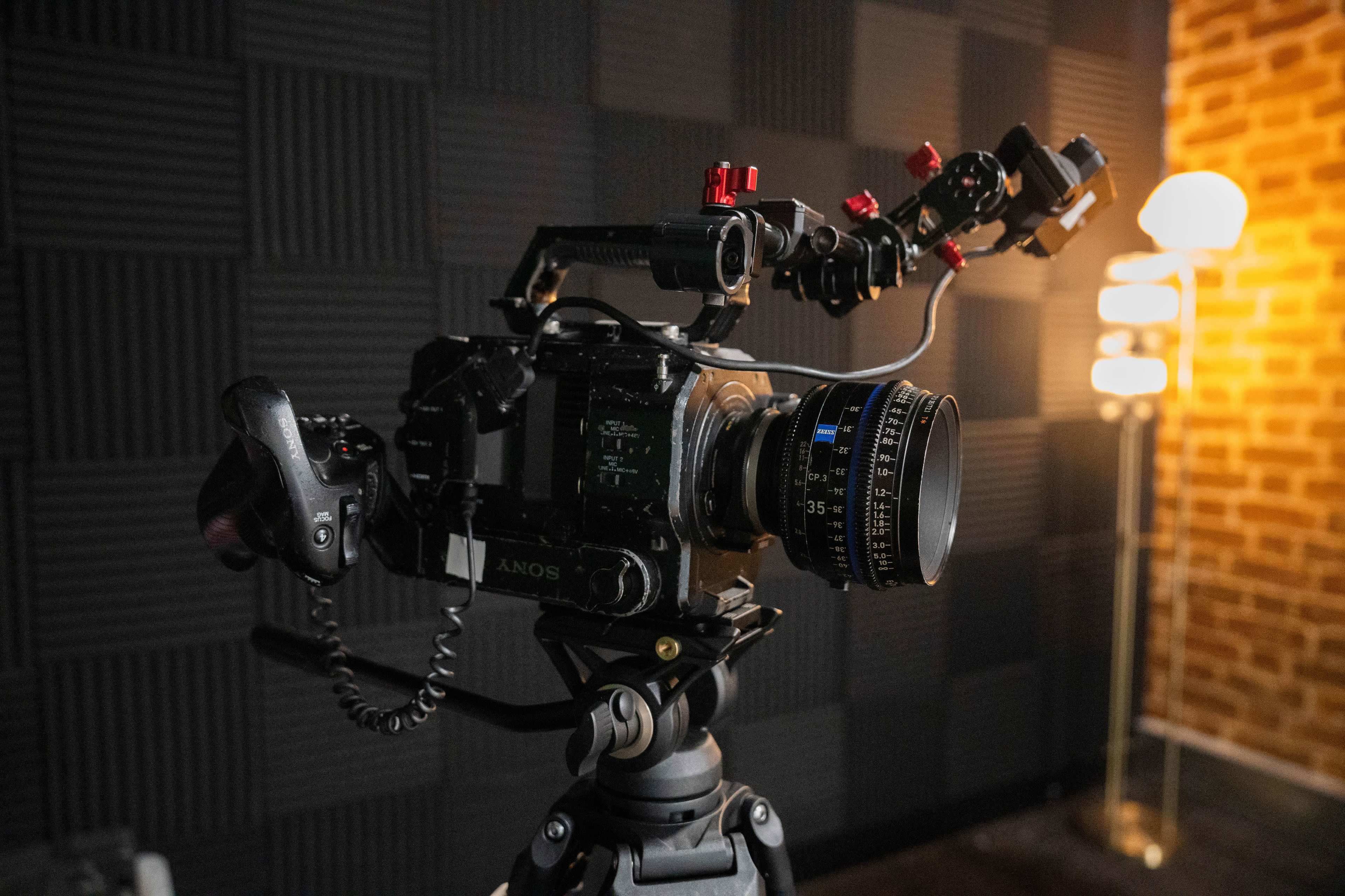 A camera is positioned on a tripod with a lens attached, set against a textured wall and a lit lamp in the background.