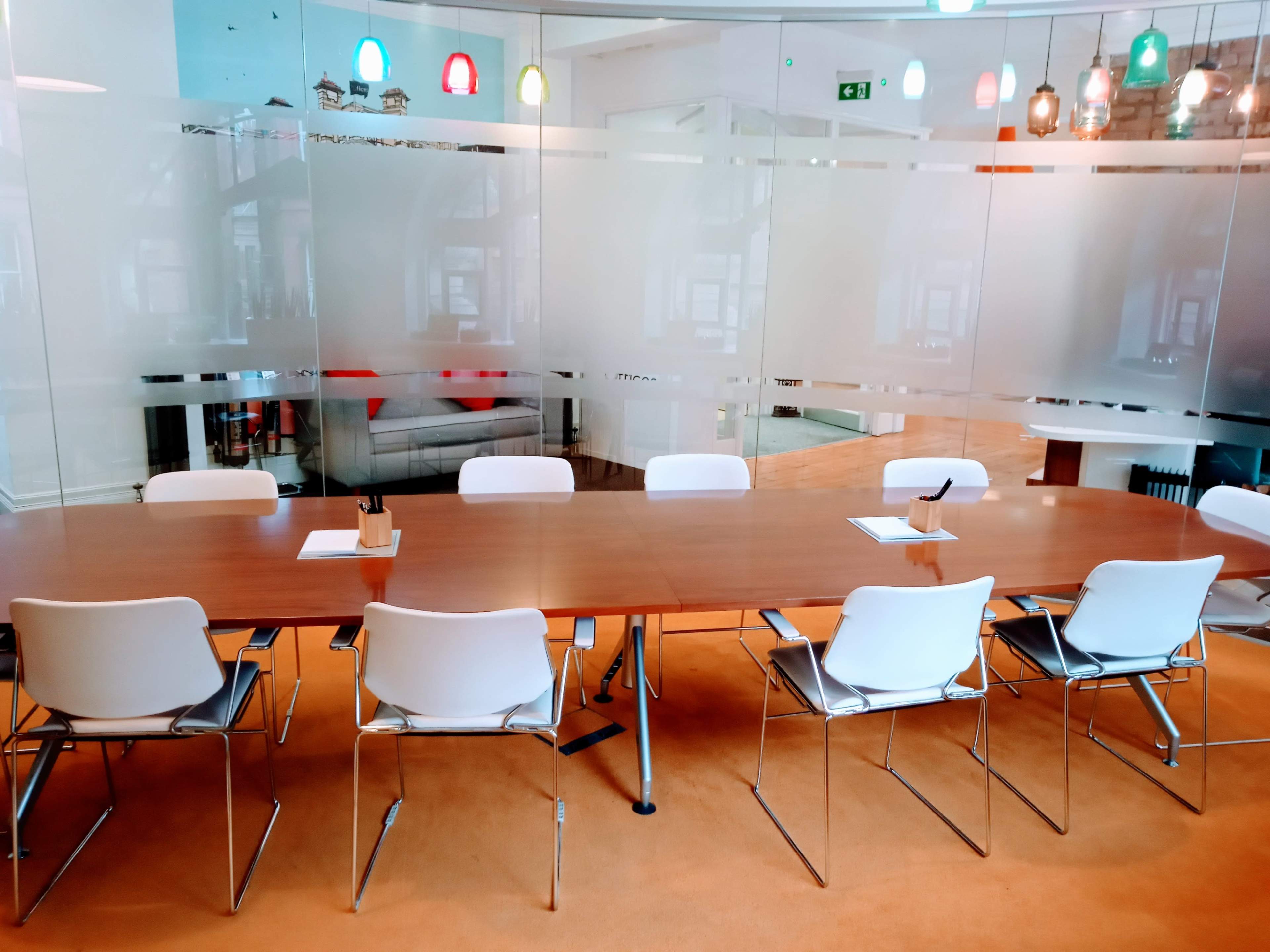 A long wooden conference table surrounded by white chairs is situated in a modern meeting room with glass partitions and colorful hanging lights.