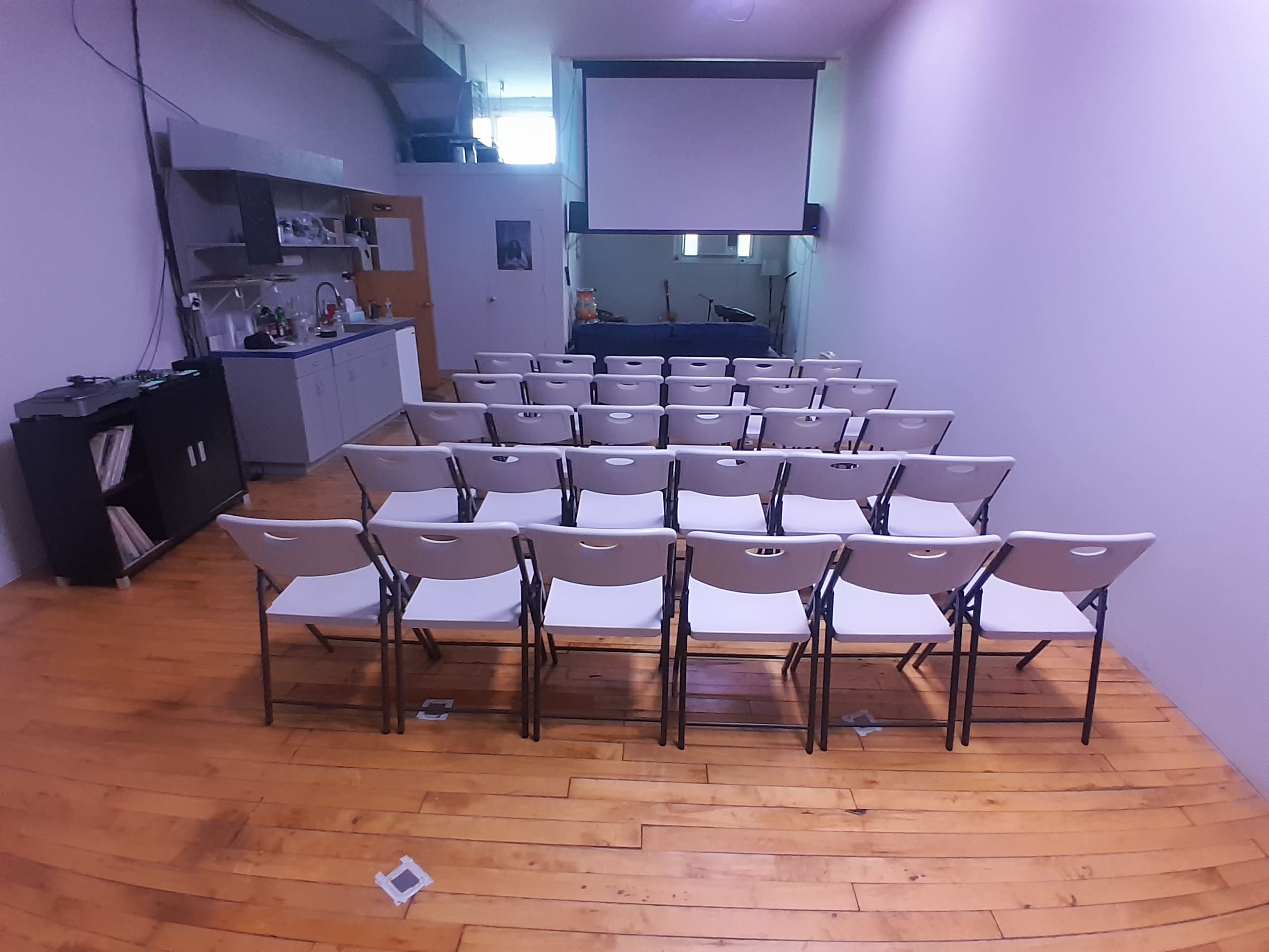 The image shows a room with a row of white folding chairs arranged in neat rows facing a large blank screen at the front, with a kitchenette area visible to the left.