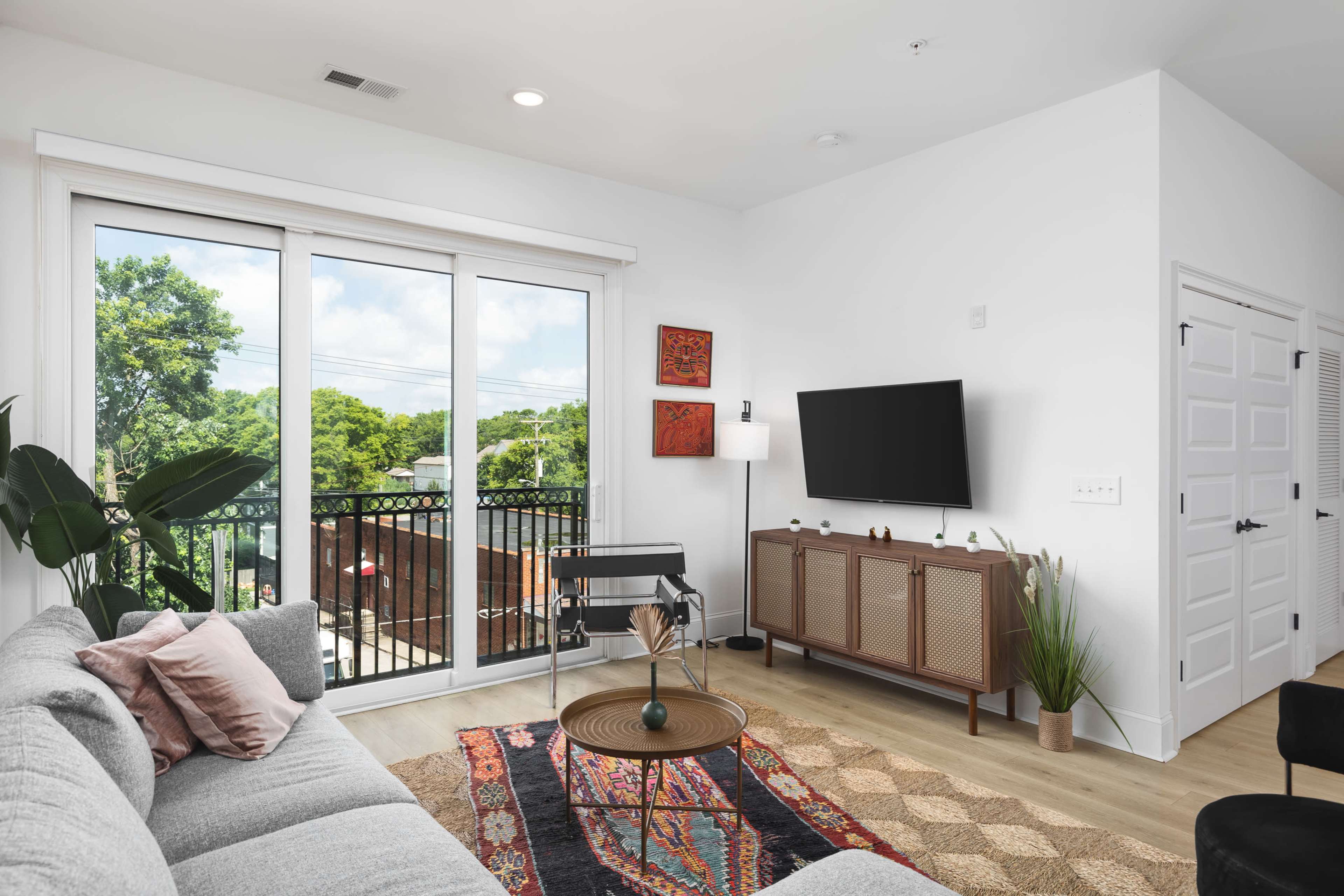 A modern living room features a gray sofa, a coffee table, a television mounted on the wall, and large windows that overlook a balcony filled with greenery.