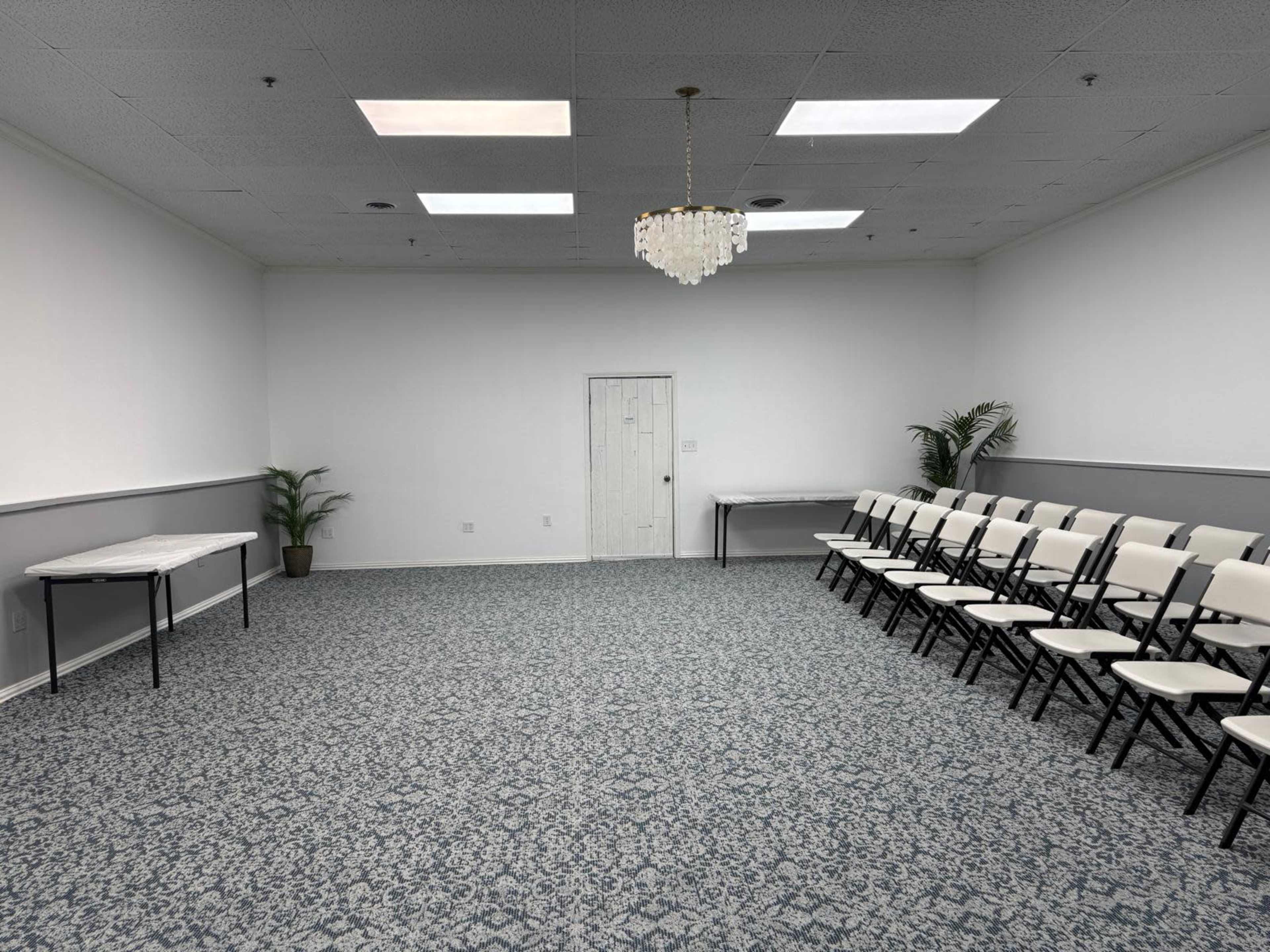 The image shows a spacious, empty room with a patterned carpet, a chandelier, and rows of white folding chairs facing a blank wall.