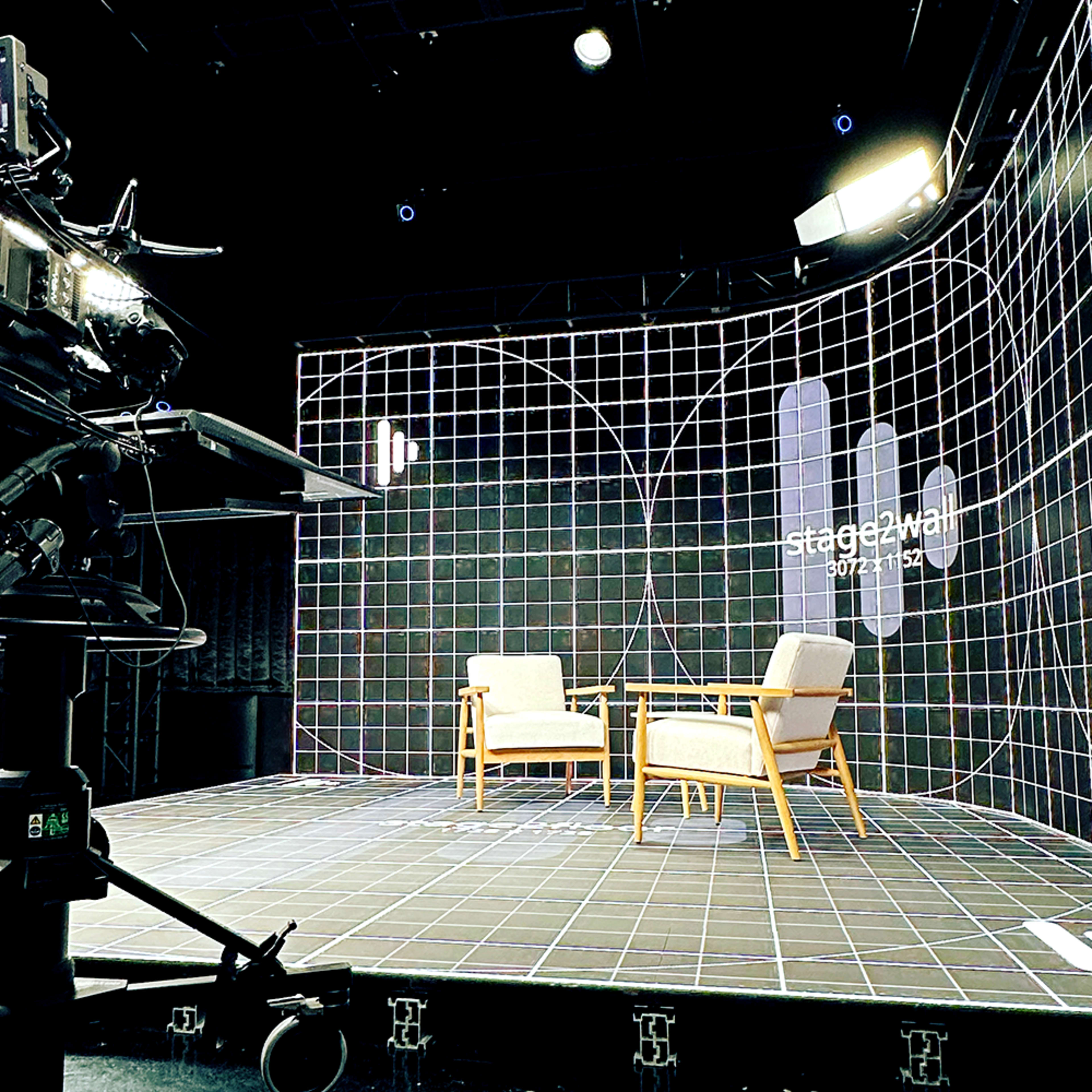 A television studio features two wooden chairs set on a grid-patterned stage backdrop with lighting equipment visible in the foreground.