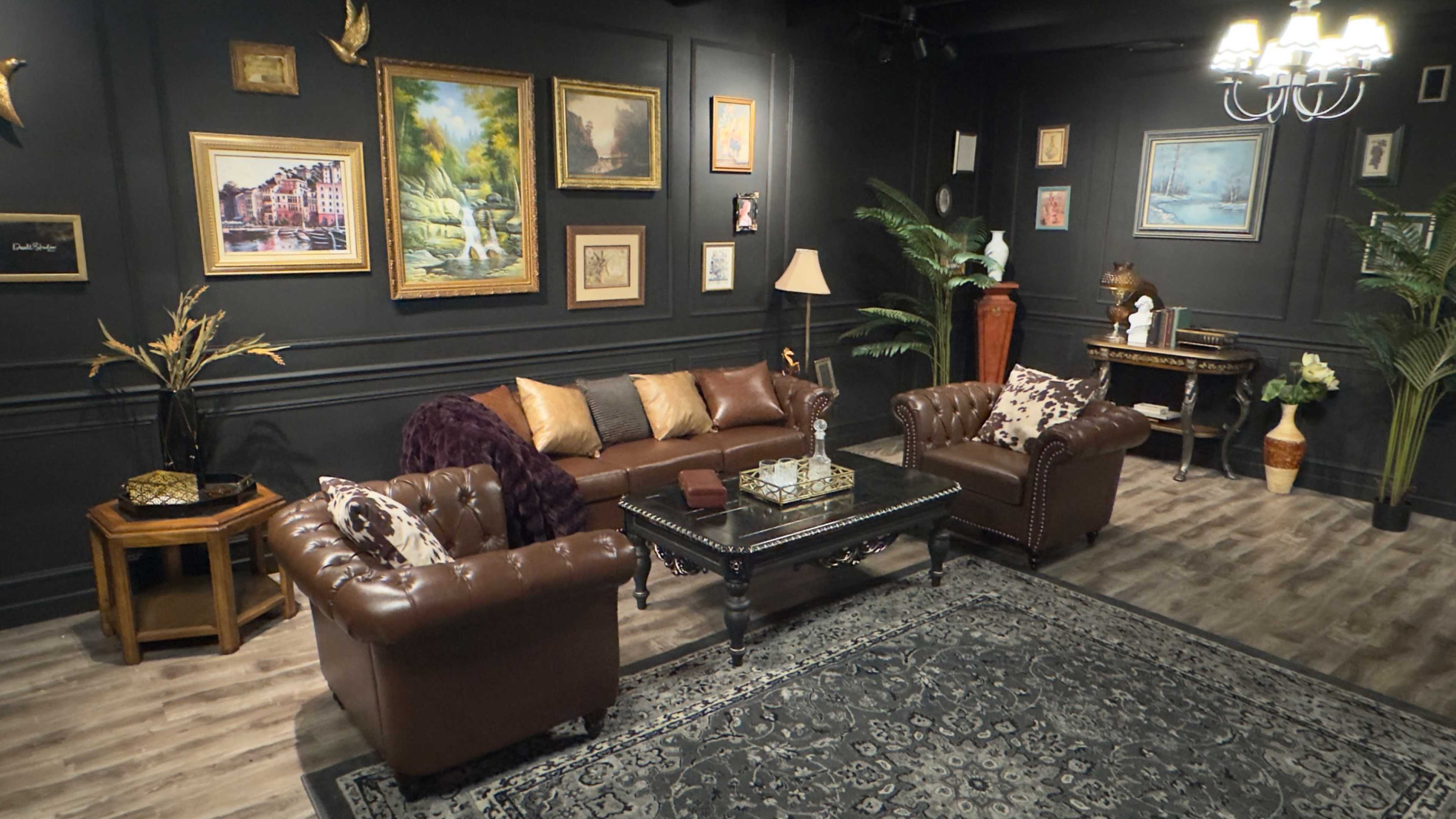 The image features a cozy living room with dark walls, a leather sofa, two armchairs, various framed paintings, and decorative plants.