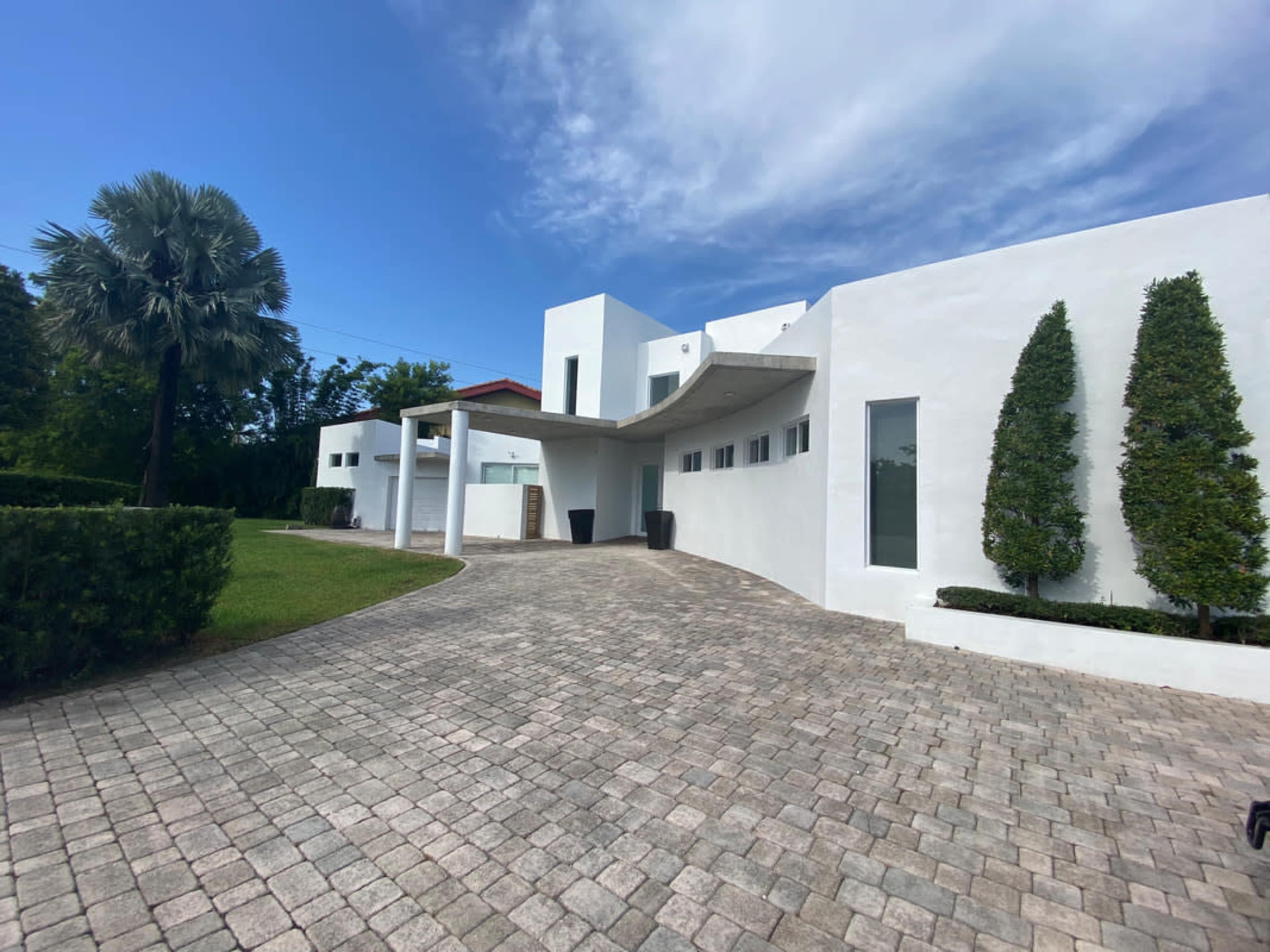 The image shows a modern white house with a landscaped driveway, flanked by palm trees and vertical shrubs under a partly cloudy sky.