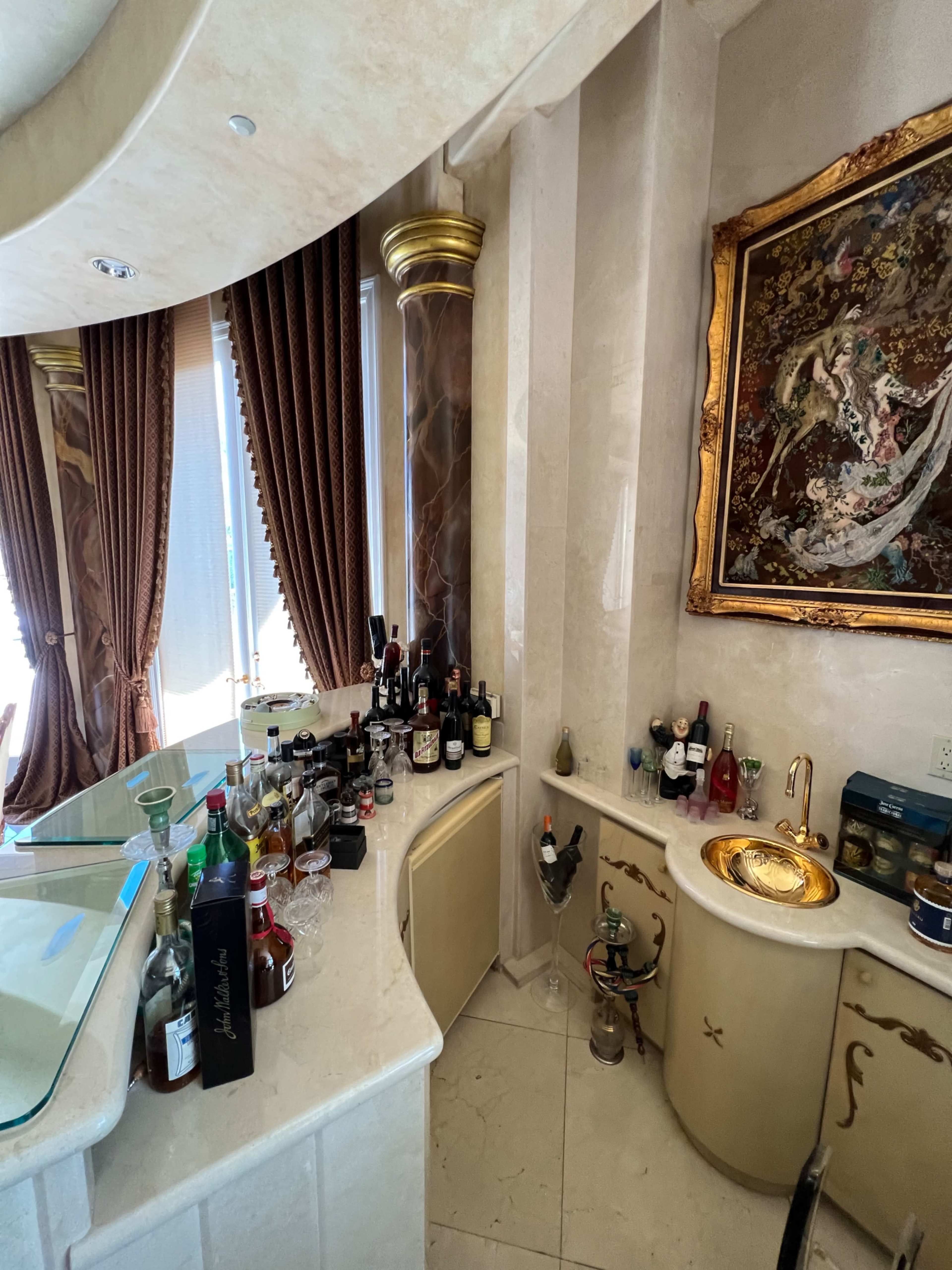 A well-organized bar area with a variety of bottles, a sink, and an ornate framed artwork in a luxurious setting.