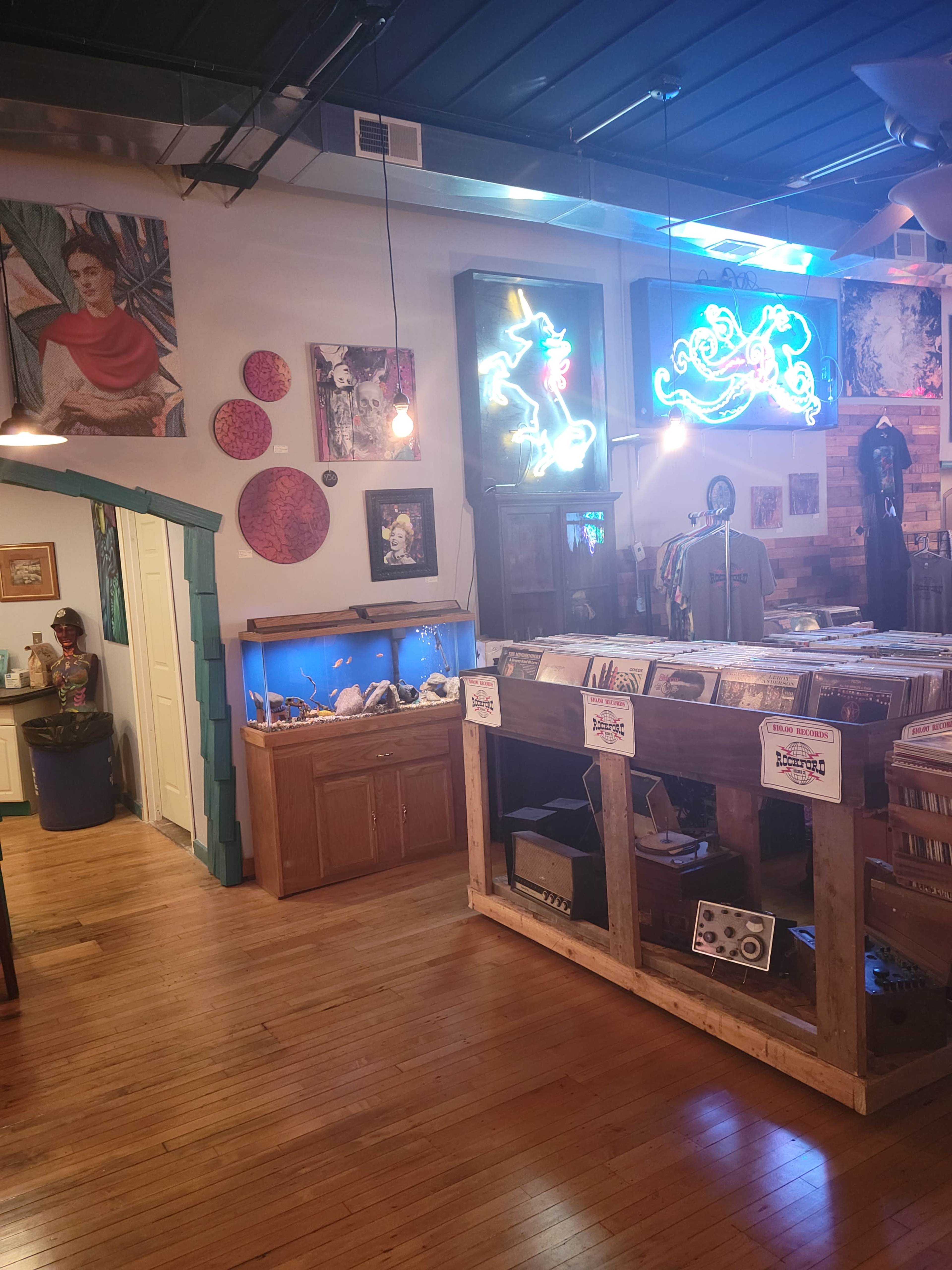 The image shows an eclectic interior of a shop featuring a wooden display counter, vinyl records, art on the walls, and a neon sign.