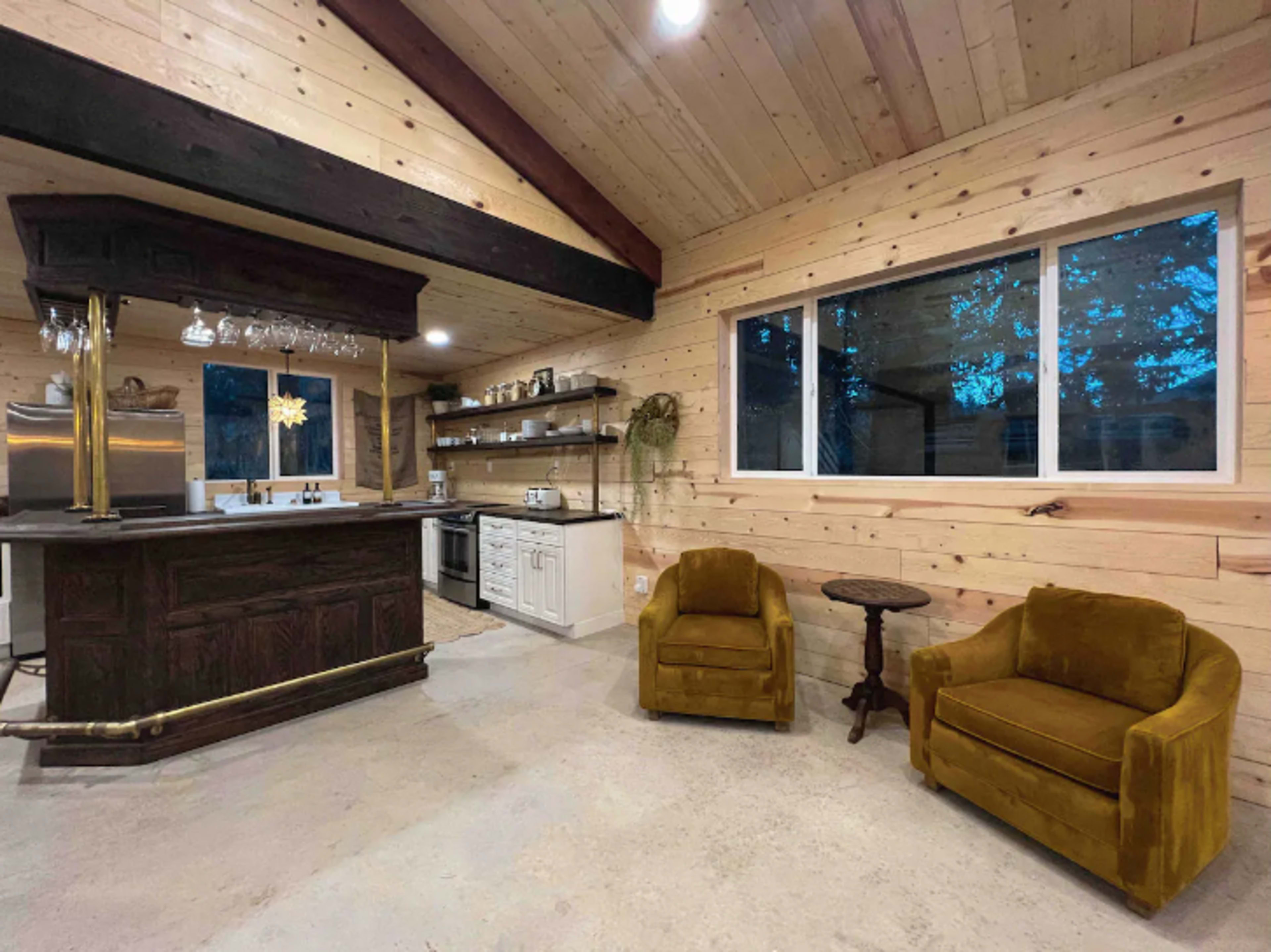 The image shows a rustic kitchen and living area featuring wooden walls, a bar with a hanging glass rack, and two yellow chairs beside a small table.